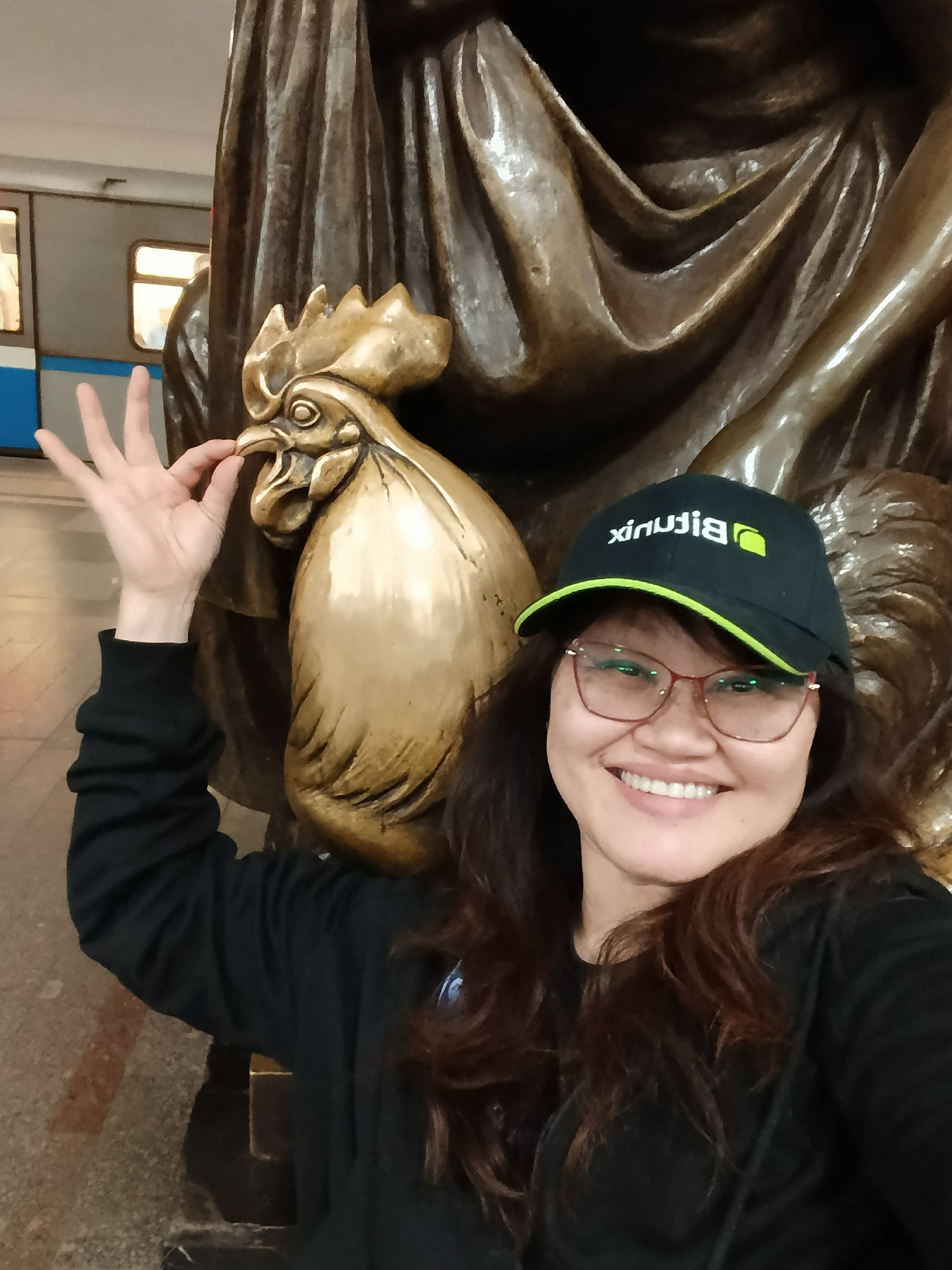 A woman is posing for a photo, smiling brightly, standing next to a large golden-bronze statue of a rooster. She is wearing a black long-sleeved top, glasses with red frames, and a black baseball cap with a green brim. The cap features a white logo and text that appears to be "Bitrix24" in reverse, indicating a selfie.
Her left hand is positioned to make an "OK" gesture, with her thumb and index finger seemingly "feeding" or touching the beak of the rooster statue. The rooster has a prominent comb and wattle, characteristic of the animal. Behind the rooster and the woman, a larger, darker bronze statue or architectural element with draped folds is visible.
In the background, to the left of the woman's head, a subway train with distinct blue and white coloring and illuminated windows can be seen, strongly suggesting the setting is a subway station platform. The overall lighting indicates an indoor, well-lit environment. The woman's broad smile conveys a sense of happiness or amusement.