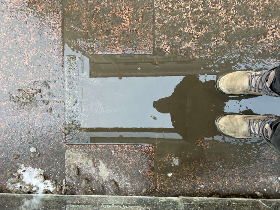 The image depicts an overhead view of a wet granite sidewalk in Saint Petersburg, Russia. In the lower portion of the frame, a person's feet are visible, clad in light brown, lace-up boots with dark laces and substantial soles, atop dark trousers. A large, rectangular puddle of murky water occupies the central and right areas of the foreground. The surface of the puddle reflects the blurred, dark silhouette of the photographer holding a mobile device, along with indistinct architectural elements of a building, including vertical lines suggesting windows or columns, and an overcast sky. The paving consists of reddish-brown granite tiles, extensively wet and discolored in areas, with scattered patches of dirty, partially melted snow and debris present, notably on the left and top-left. A vertical, dark, wet structural element, possibly a curb or railing, runs along the far-left edge of the frame.
