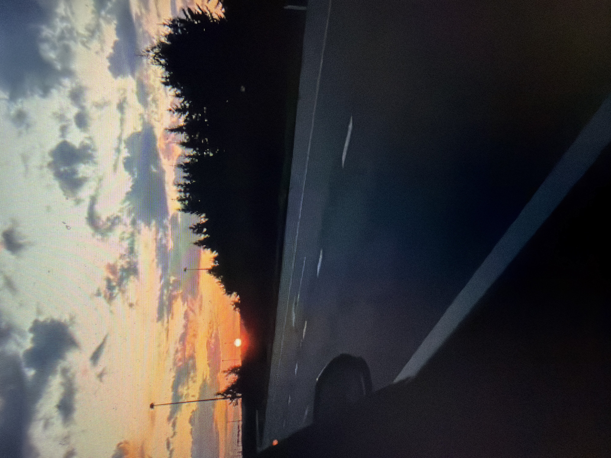 The image shows a highway at sunset. The sky is filled with colorful clouds, ranging from orange and pink to gray and blue. The sun is setting on the horizon, casting a warm glow on the scene. Silhouetted trees line the side of the highway, and a street light can be seen in the distance. The road is asphalt and has white lane markings. A car is partially visible in the foreground on the left, with its side mirror reflecting the sky. The overall mood of the image is peaceful and serene.