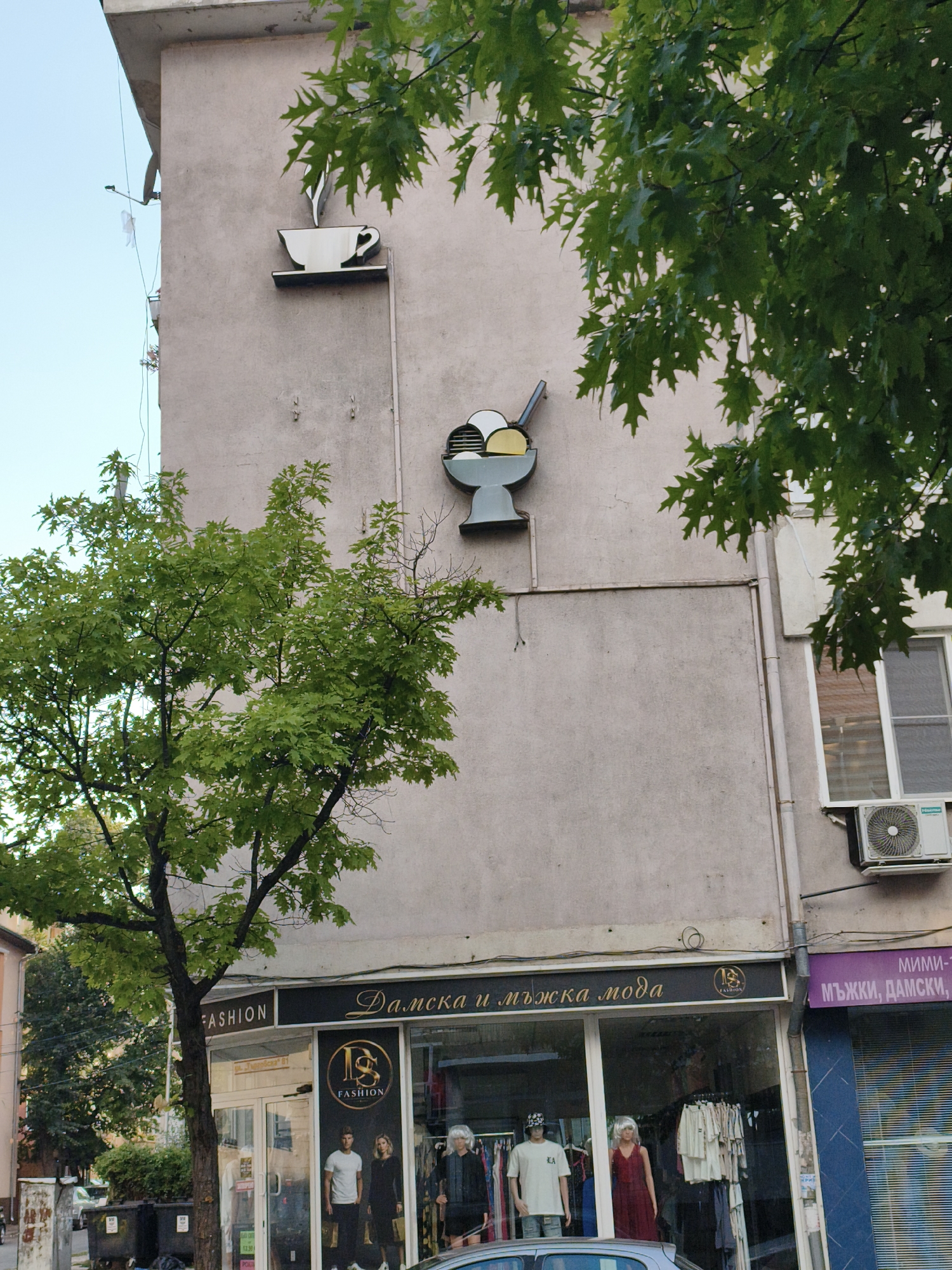 The image depicts the exterior of a building in Lovech, Bulgaria, with a storefront on the ground floor. The building's facade is a plain, light-colored concrete. Mounted on the upper part of the building are two decorative signs: one is a white silhouette of a steaming cup on a saucer, and the other is a blue and grey representation of an ice cream sundae. Green foliage from a tree in the foreground partially obscures the view.
The storefront below the signs is for a clothing store labeled "FASHION" and "Дамска и мъжка мода" (Women's and Men's Fashion). The display windows showcase mannequins dressed in clothing, as well as racks of garments. To the right of this storefront, another shop window is partially visible, with a purple sign reading "МИМИ- Т" and "МЪЖКИ, ДАМСКИ" (Men's, Women's). An air conditioning unit is mounted on the wall above this second shop. The environment appears to be a street scene with typical urban elements like trash bins and a parked car in the foreground. The sky is clear and blue.