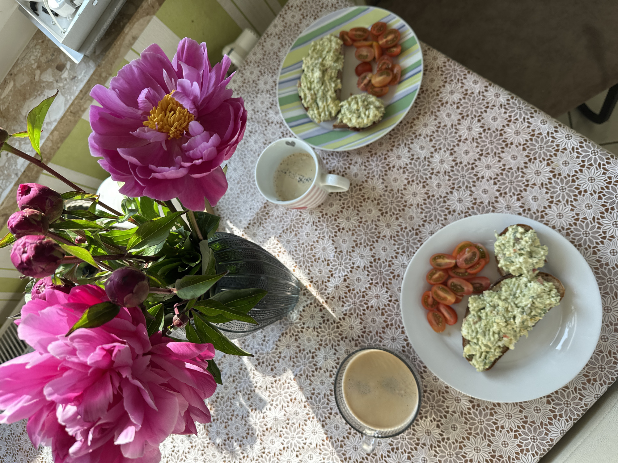 A bright, overhead shot shows a table setting for breakfast in Fredenbeck, Germany. The table is covered with a white lace tablecloth featuring a floral pattern. On the table, there are two plates of food, two cups of coffee, and a vase of pink peonies. One plate holds two pieces of dark bread topped with a green spread and sliced cherry tomatoes. The other plate has a similar spread on bread and a pile of cherry tomatoes. One cup of coffee is in a clear glass, while the other is in a white mug with pink stripes. The vase of peonies, a vibrant pink with yellow centers, sits to the left of the plates and cups. Sunlight streams in from the left, casting shadows across the tablecloth. The background is blurred, but hints of green and beige walls and what appears to be a radiator are visible.