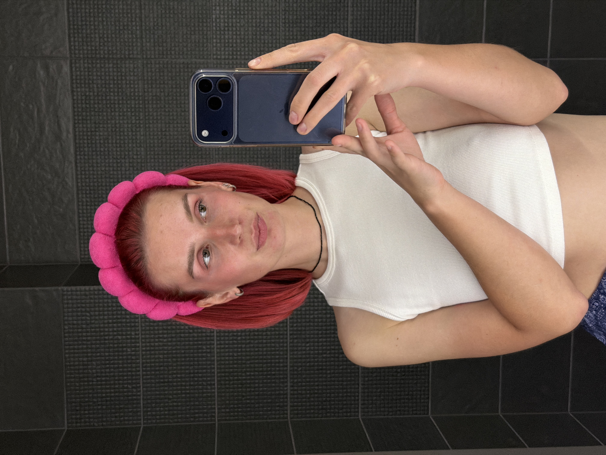 A person is taking a selfie with a smartphone. The person has bright red hair, is wearing a white ribbed tank top, and a pink puffy headband. They are also wearing a black beaded necklace and have silver hoop earrings. The background appears to be a tiled wall. The person is holding a dark blue smartphone in their hands, pointing it towards themselves to take the picture. Their expression is neutral.