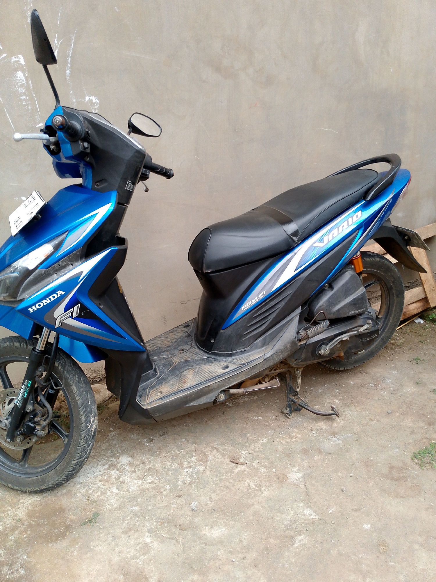 A blue and black Honda Vario scooter is parked on a concrete surface in front of a textured, beige wall. The scooter has a black seat, a kickstand extended, and a license plate is attached to the front. The word "HONDA" is visible on the front fairing, and "VARIO" is on the side panel. The ground is dirty with some grass patches and debris. The scene appears to be outdoors, possibly in a residential or informal urban area in Babadan, Indonesia. No people are present in the image.