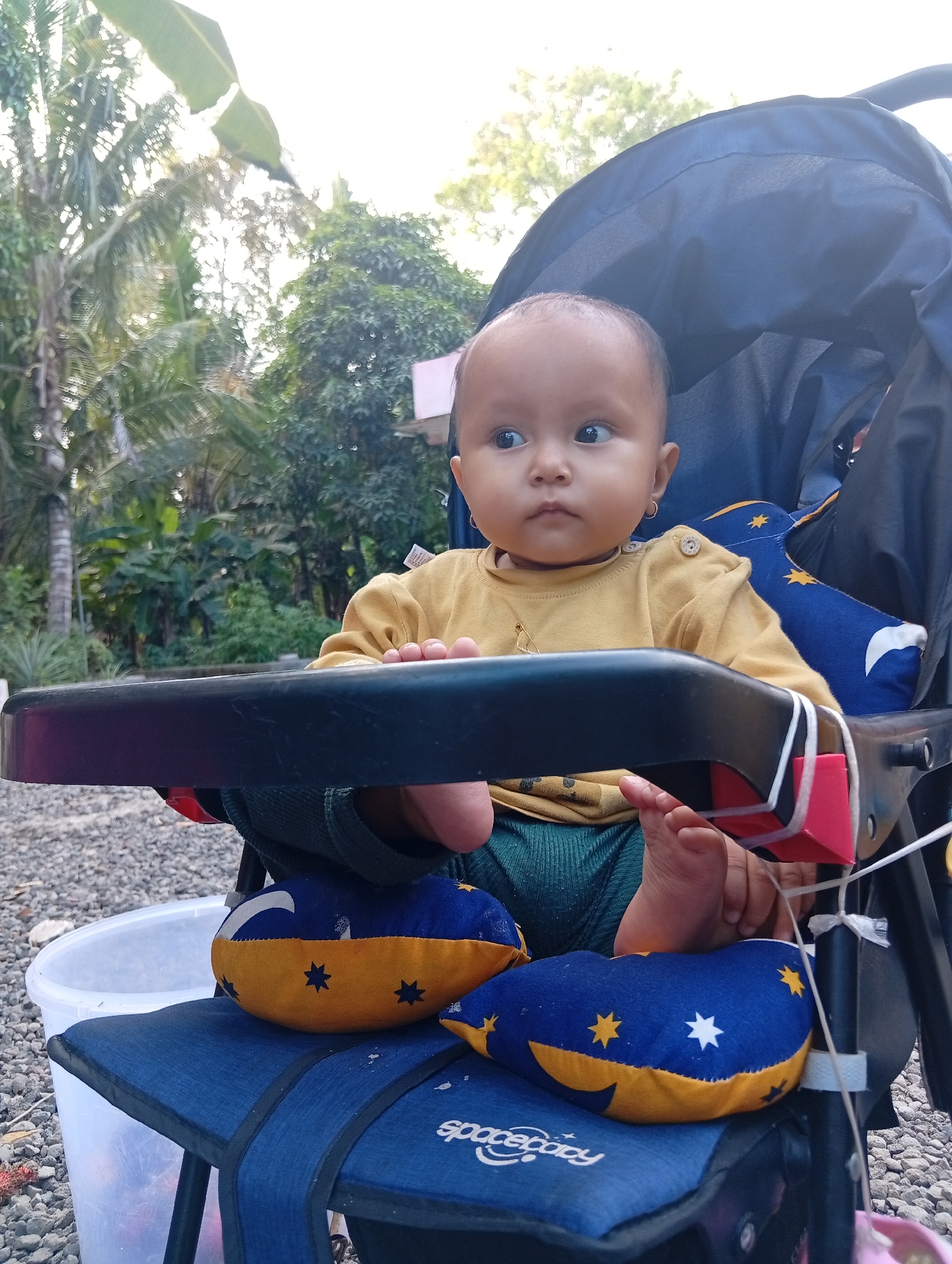 A young baby sits in a blue stroller, facing the camera, with a blurred outdoor background of trees and foliage, suggestive of a tropical or humid climate, possibly in Miritpetikusan, Indonesia. The baby, with dark hair and brown eyes, wears a mustard-yellow long-sleeved shirt and dark green pants. A tray is in front of the baby, with a small pink object attached. The stroller has blue fabric with yellow and blue star and moon patterns on its cushions and a prominent dark blue canopy. A clear plastic bucket is visible to the left of the stroller. The ground is covered with small gray stones. The lighting suggests it is daytime, possibly late afternoon, with sunlight filtering through the trees.