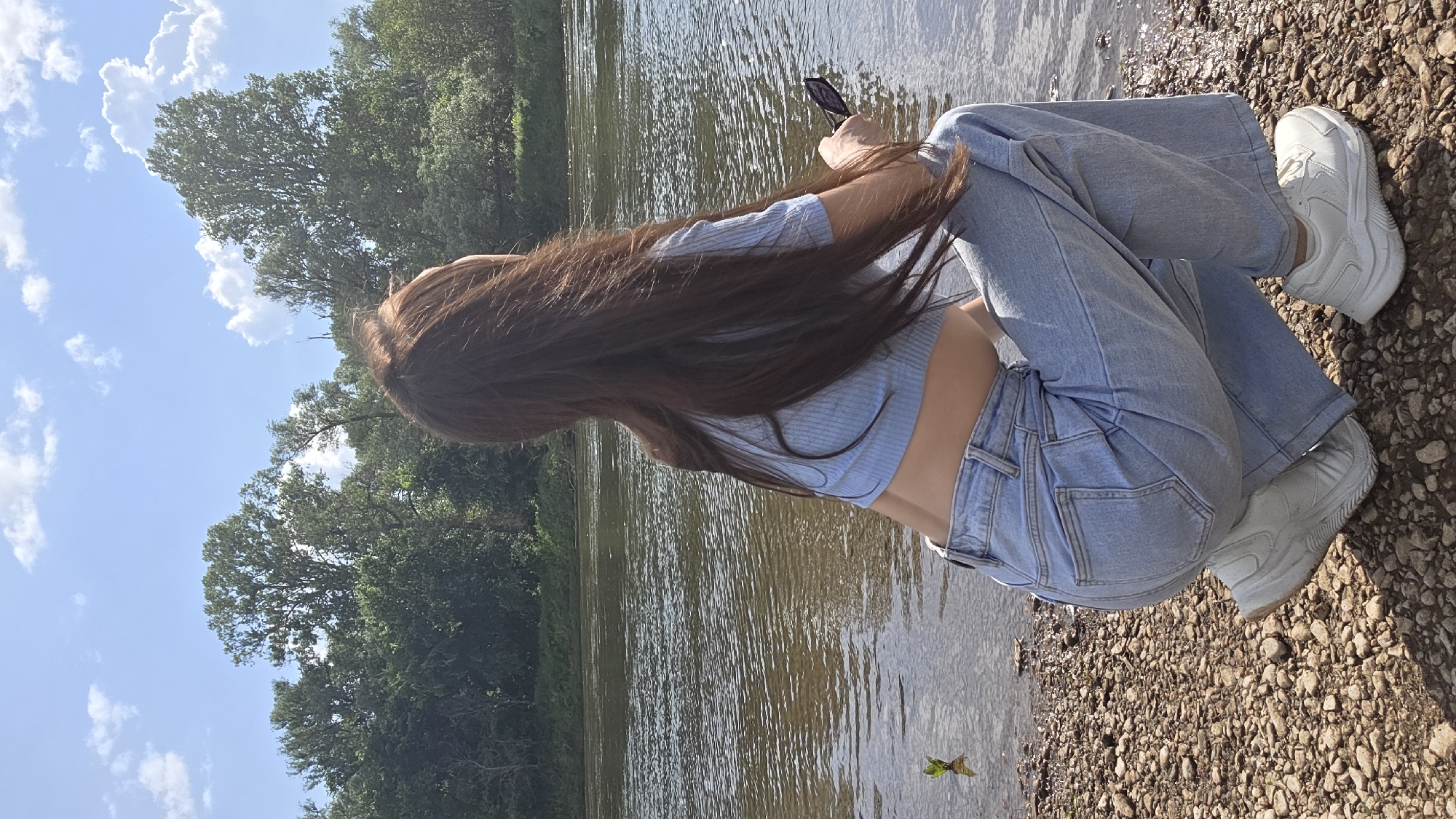 The image shows a person from the back, sitting on a rocky shore next to a body of water. The person has long, dark hair that extends over their shoulders. They are wearing a light blue crop top and light blue baggy jeans. On their feet are white platform sneakers. In their right hand, they are holding a dark-colored smartphone. The background consists of trees and a bright blue sky with scattered white clouds. The water appears to be a river or lake, with ripples on its surface. The sun is shining, indicating it is daytime. The overall scene is outdoors, likely in a natural setting. The person's posture and focus on their phone suggest they are engaged with the device. The lighting suggests a sunny day.