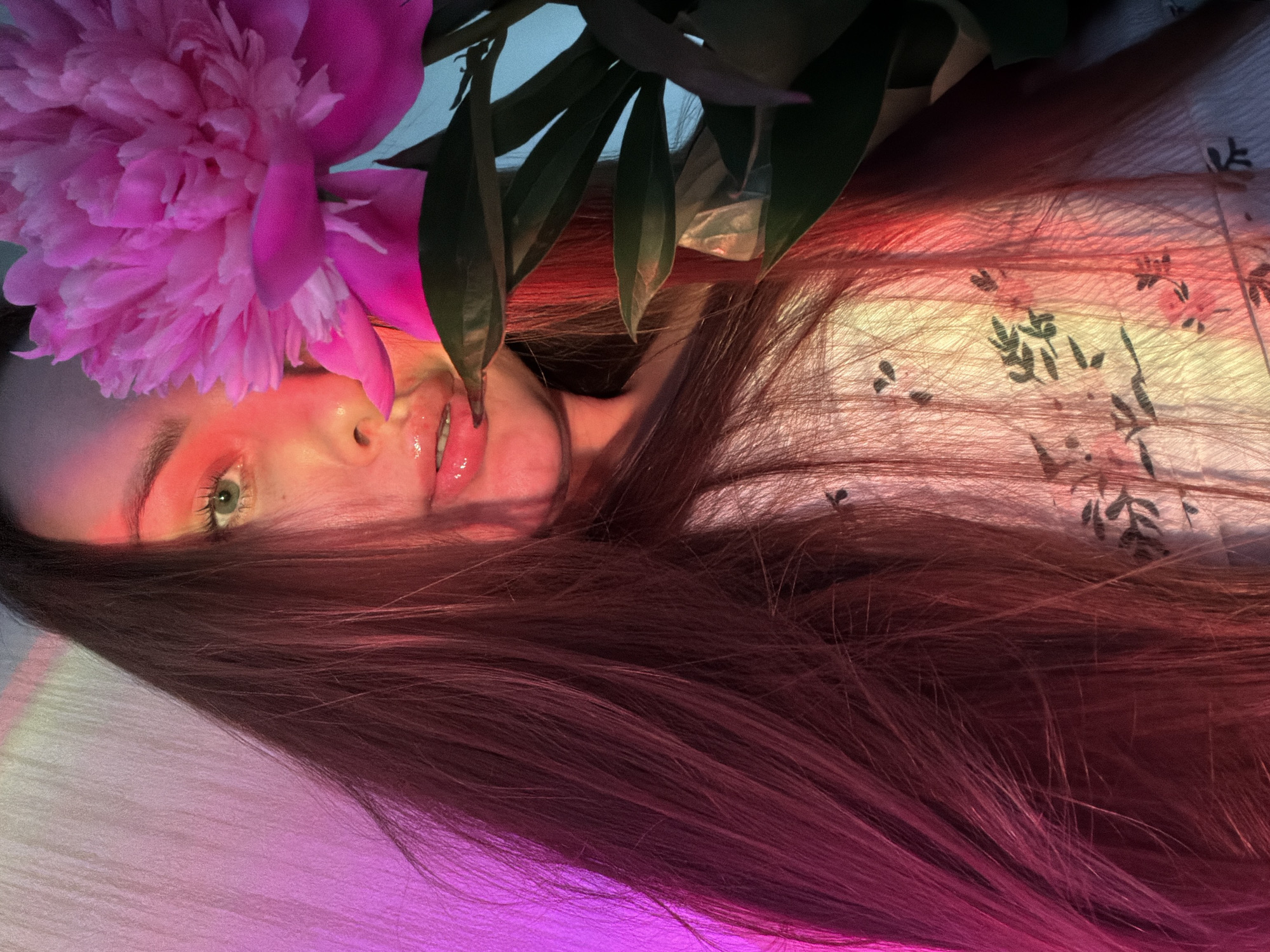 The image shows a close-up, low-angle portrait of a woman with long, dark hair partially obscured by a large pink peony flower. She has green eyes, glossy pink lips, and is wearing a white top with a floral pattern. The scene is illuminated by pink and yellow lighting, creating a dramatic effect on her face and hair. The background appears to be a textured white surface. There is no identifiable information about the location in Chaikovsky, Russia.