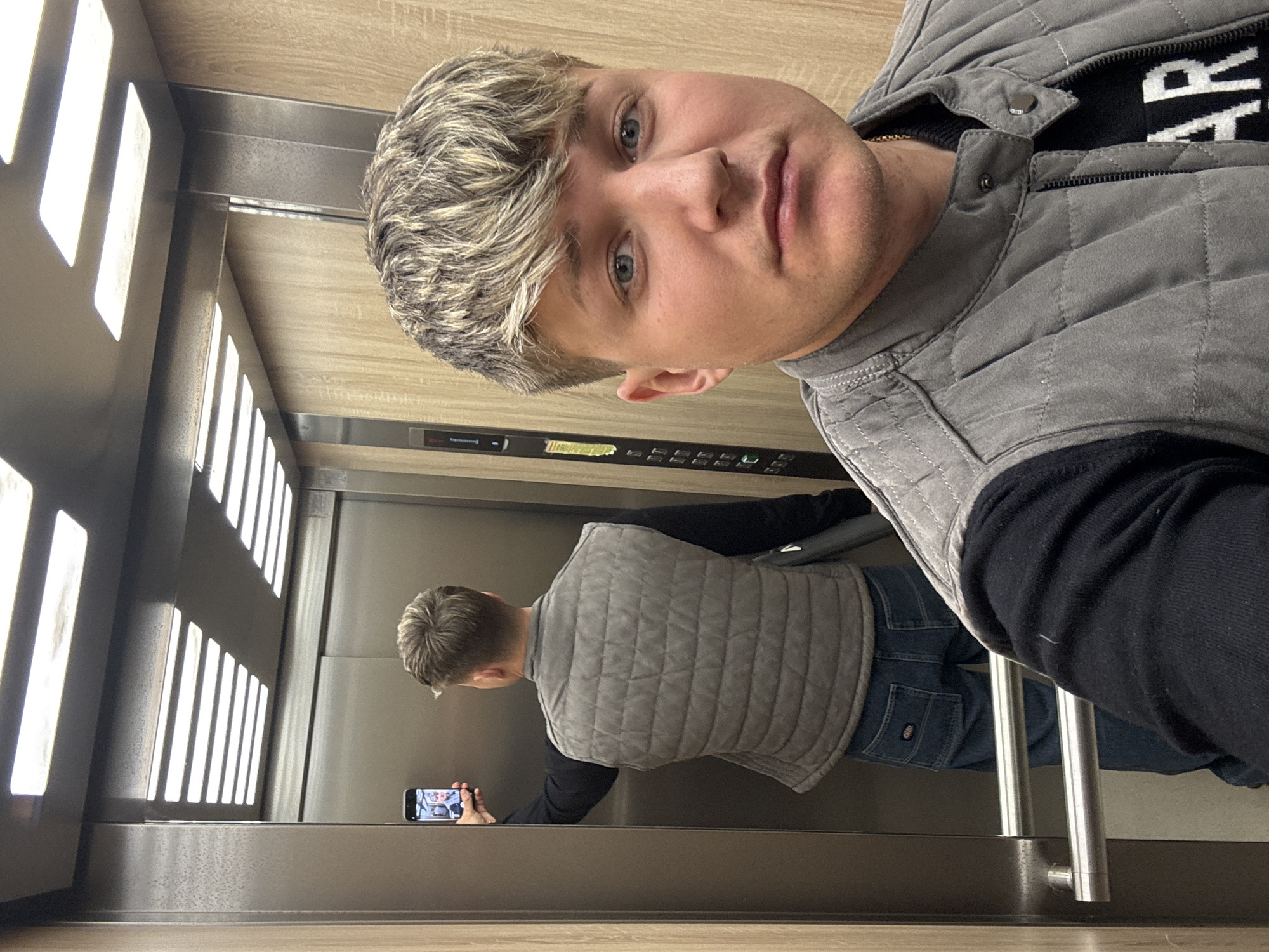 A young, fair-skinned man with blond hair takes a selfie in a modern elevator. He wears a gray quilted vest over a black long-sleeved shirt and blue jeans. His reflection is visible in the mirror, where he is holding a smartphone, seemingly taking the picture. The elevator walls are paneled with wood, and the lighting is provided by recessed rectangular lights. A metal handrail is visible. The location is unknown, the prompt has specified the city of Plovdiv, Bulgaria, although there is no way to confirm this location from this image alone.