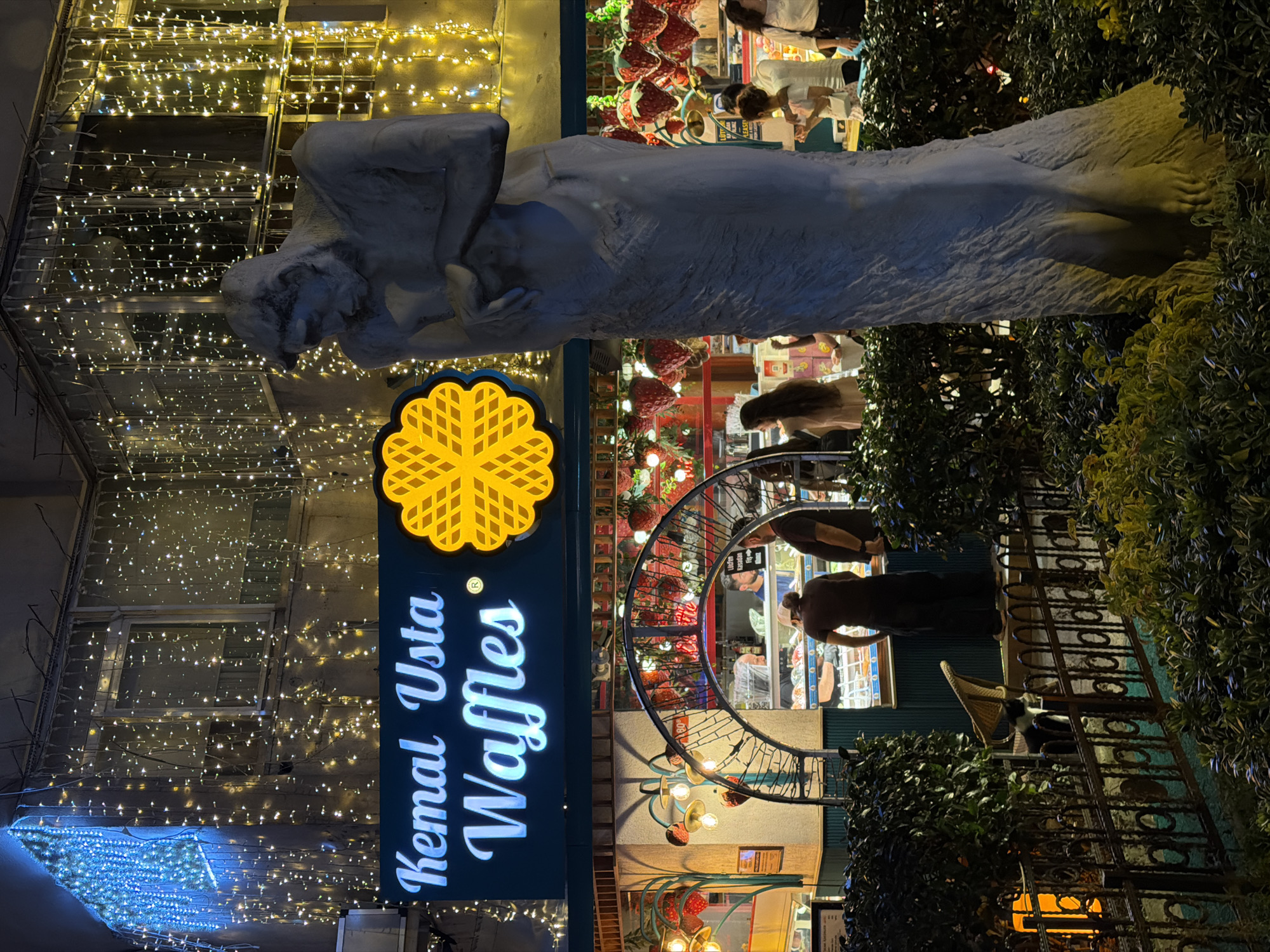 The image captures a nighttime scene outside "Kemal Usta Waffles" in Kadıköy, Turkey. The storefront is adorned with cascading strings of white and blue fairy lights, creating a festive atmosphere. In the foreground, a tall, grey statue of a person stands to the right, partially obscuring the waffle shop. The shop itself features an arched entryway with decorative lights and displays of strawberries. Inside, several people are visible, including staff serving customers. A black and white cat is visible on a fence in the lower left portion of the image, amidst lush green foliage. The overall impression is that of a vibrant street-level establishment in a lively urban environment.