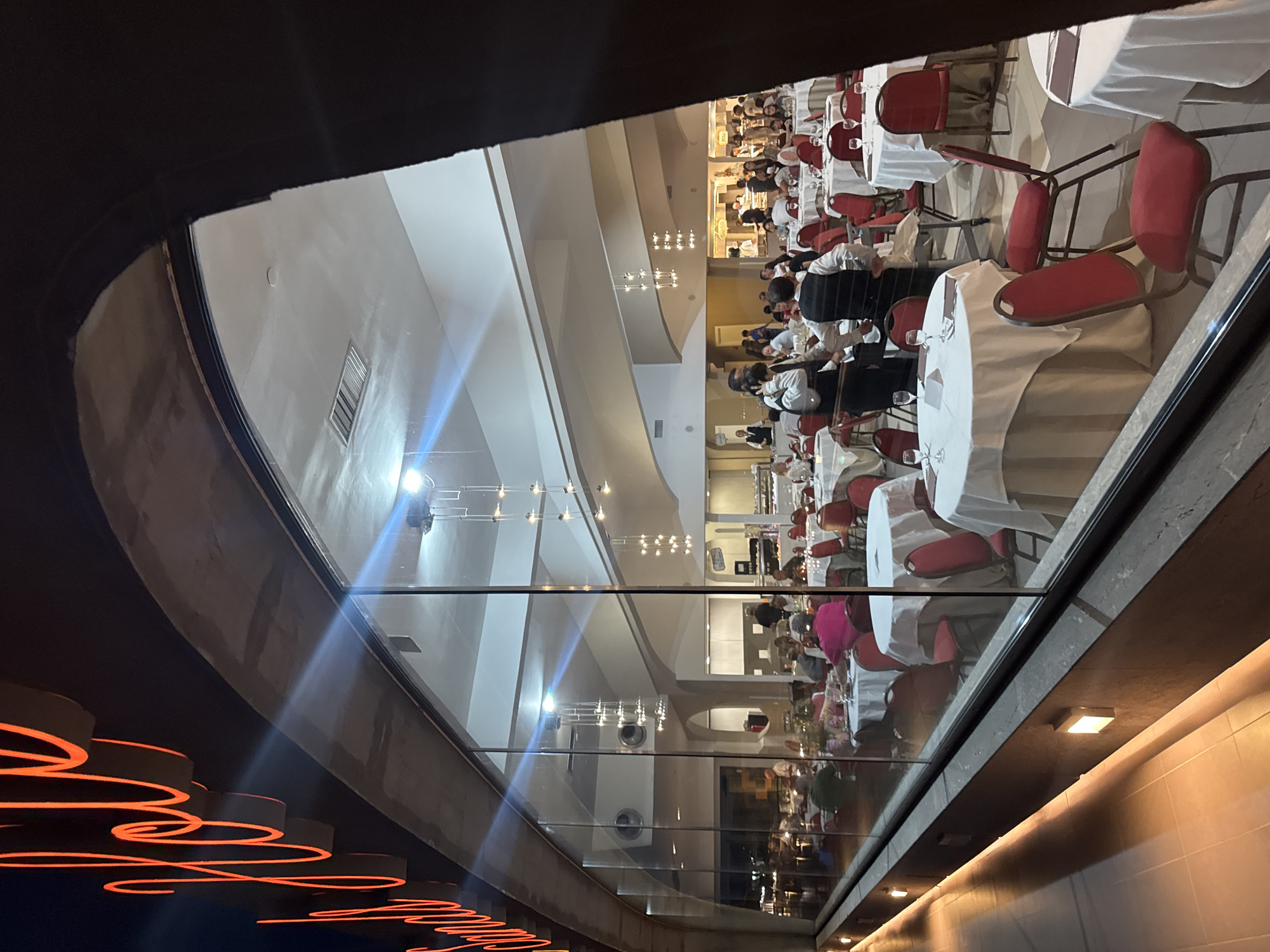 The image shows a large, well-lit restaurant in Capaci, Italy, viewed from the outside through a large glass window. The restaurant appears to be busy, with many tables set for dining and patrons seated. Waitstaff in uniforms are visible, attending to tables and moving through the dining area. The interior features high ceilings, modern light fixtures, and a buffet or serving area in the background. The exterior of the building displays a large, illuminated orange sign with cursive lettering. The foreground shows a walkway with accent lighting.