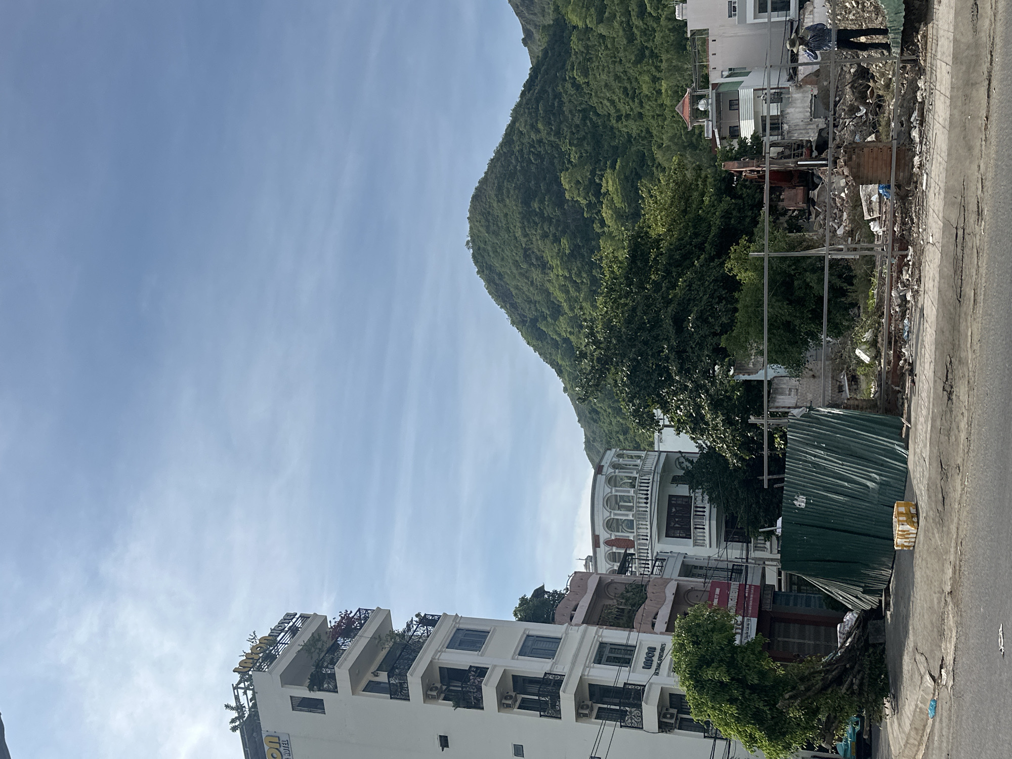 The image shows a street scene in Bac Nha Trang Ward, Vietnam, on a clear day. In the foreground, there are several buildings, including a white multi-story apartment building with balconies and air conditioning units, and another building with a more ornate facade and arched windows. A green metal sheet fence is erected along the sidewalk, partially obscuring a construction site with rubble and a red excavator. In the background, a lush green mountain slopes upwards into a bright blue sky with wispy clouds.
There are a couple of people visible in the background, one wearing a hat and standing near the construction site, and another person or two further in the distance near the buildings. The scene appears to be a mix of ongoing construction or demolition and established residential or commercial buildings. The time of day is likely midday or early afternoon, judging by the bright sunlight and clear sky. The location is suggested by the text "LIVON APARTMENTS" visible on one of the buildings and the overall architectural style. A yellow sign with red text is also visible on one of the buildings.