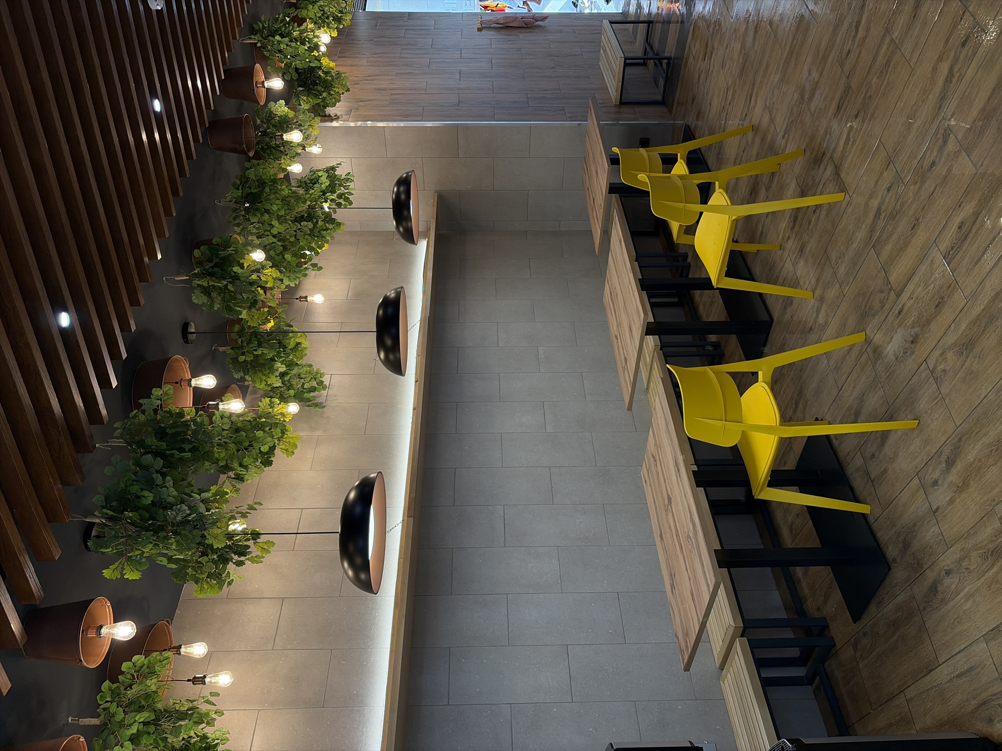 The image depicts the interior of a commercial establishment, likely a cafe or fast-casual restaurant, located in Votkinsk, Russia. The environment features a blend of modern and industrial aesthetics with natural decor elements. The left wall is constructed from light grey rectangular tiles, topped by a horizontal wooden shelf with a concealed strip light providing upward illumination. The right wall is clad in vertical wooden panels. The ceiling is characterized by dark wooden beams against a dark background, from which numerous clusters of artificial green foliage are suspended. Embedded within this foliage and hanging independently are multiple exposed Edison-style light bulbs, some of which are contained within inverted terra cotta-colored pots. Three prominent black dome-shaped pendant lights, each with a copper-toned interior, are suspended at various points above the seating area.
In the foreground and midground, three rectangular wooden tables with black metal bases are visible. Two of these tables are paired with slatted wooden benches featuring black metal frames. Three bright yellow plastic chairs are strategically placed around the tables. The floor is covered with brown wood-patterned ceramic tiles, which exhibit a wet or highly reflective appearance in several areas, particularly on the right side. No individuals are present within the scene.