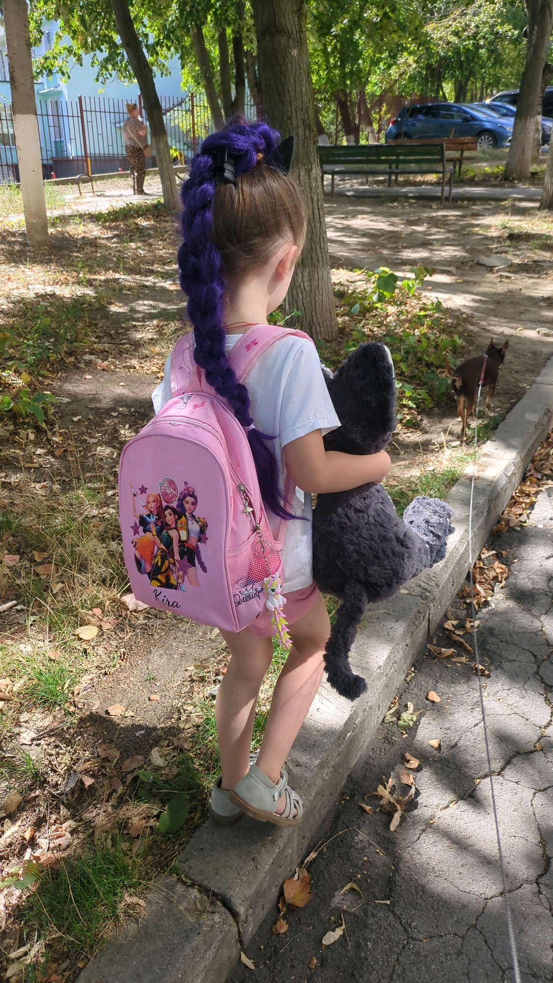 The image depicts a young girl with a long, braided purple hairstyle walking outdoors. She is wearing a white t-shirt, pink shorts, and grey sandals. The girl is carrying a pink backpack adorned with cartoon characters and the name "Kira". She is also hugging a large, dark grey stuffed animal. A small, dark brown dog is visible in the background, standing on a leash. The scene appears to be a park or a sidewalk area with trees, grass, and a concrete curb. In the distance, there are buildings and parked cars, suggesting an urban environment. The overall lighting suggests it is daytime. Without specific landmarks or identifying signage, it is not possible to definitively pinpoint the location within Chișinău, Moldova.