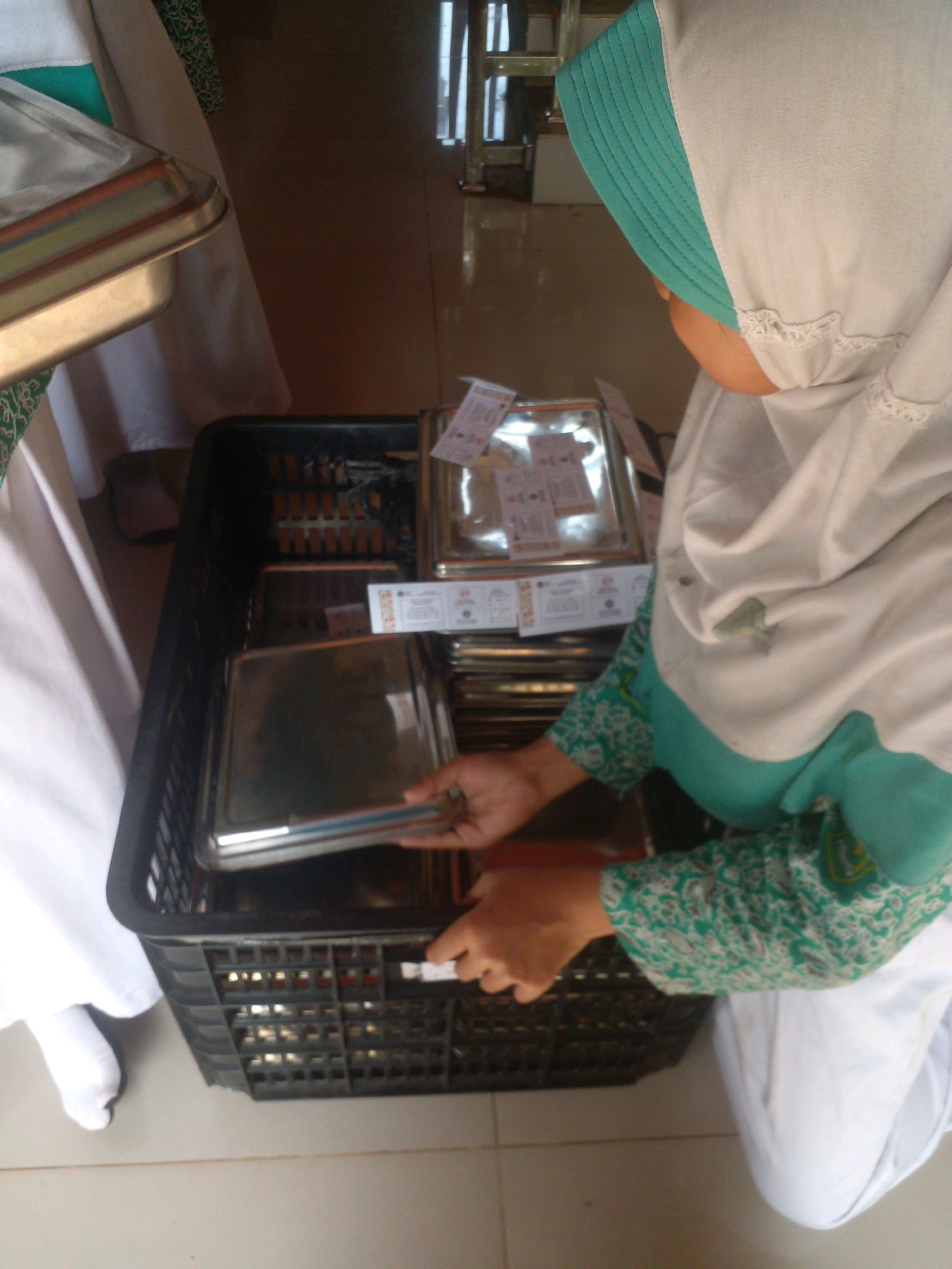 In a low-angle, close-up shot, a person wearing a white hijab and a green and white patterned garment is seen sorting metal serving dishes. The person's hands are visible as they hold and arrange the dishes inside a black plastic crate. The crate is filled with more serving dishes, some stacked neatly and others appearing slightly disarranged. In the background, the floor is visible, and parts of what appear to be other people in white garments are present. The scene is likely indoors, possibly in a catering or food service environment, given the presence of numerous serving dishes. The location in Banyuputih, Indonesia, is noted.