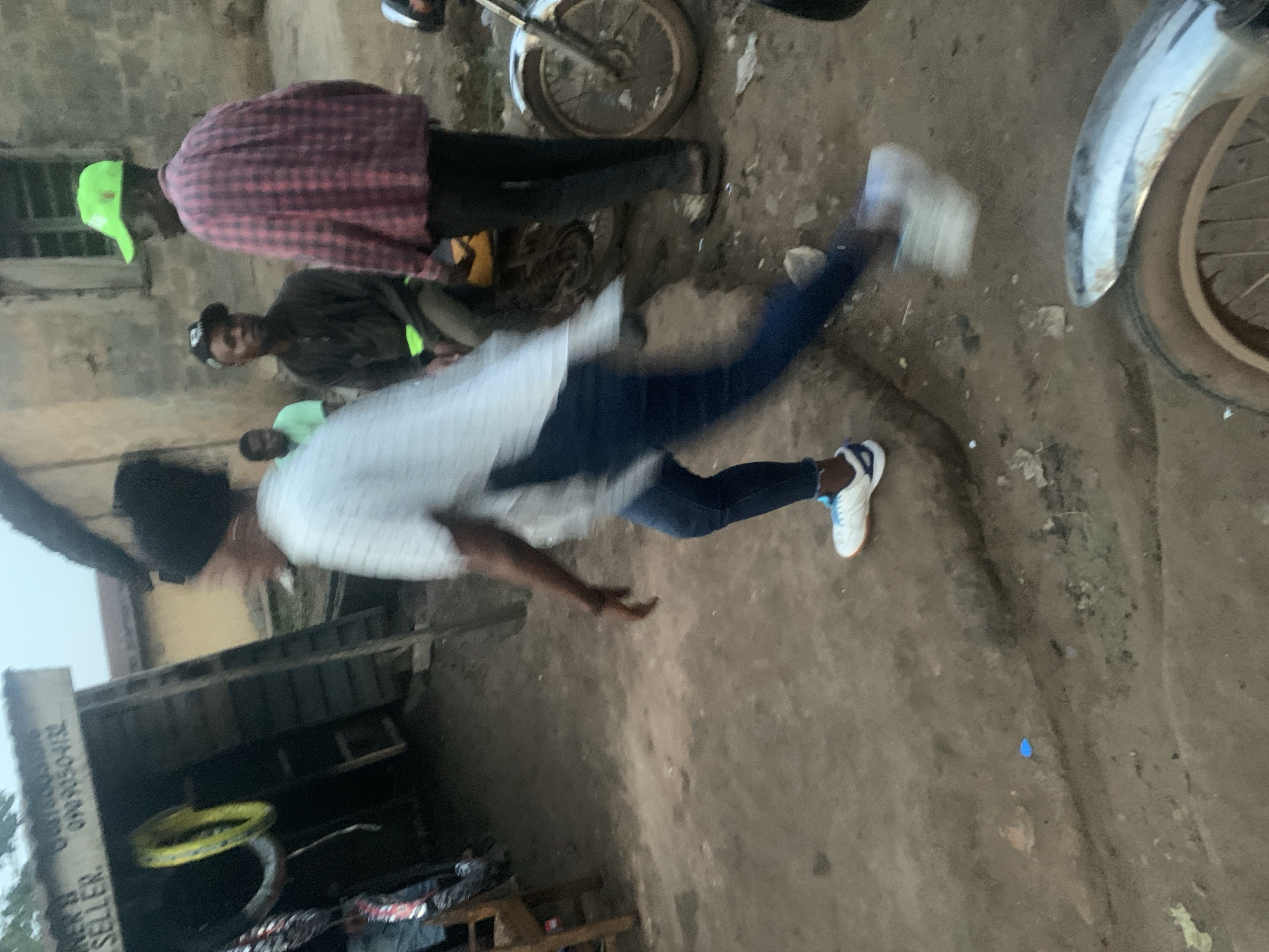 In Shagamu, Nigeria, a person wearing a white shirt and blue jeans is captured in motion, their body blurred by movement. They are running on a dirt ground in front of a structure with wooden walls and a corrugated metal roof. A sign on the structure reads "SELLER" and displays a phone number. Yellow tire rims are visible inside the structure. To the right of the runner, a person in a red and black checkered shirt and a green cap is talking to another person in a dark shirt and a black baseball cap. A motorcycle is parked to the far right of the scene, with its front wheel visible. The environment appears to be a dusty, outdoor area with some debris on the ground.