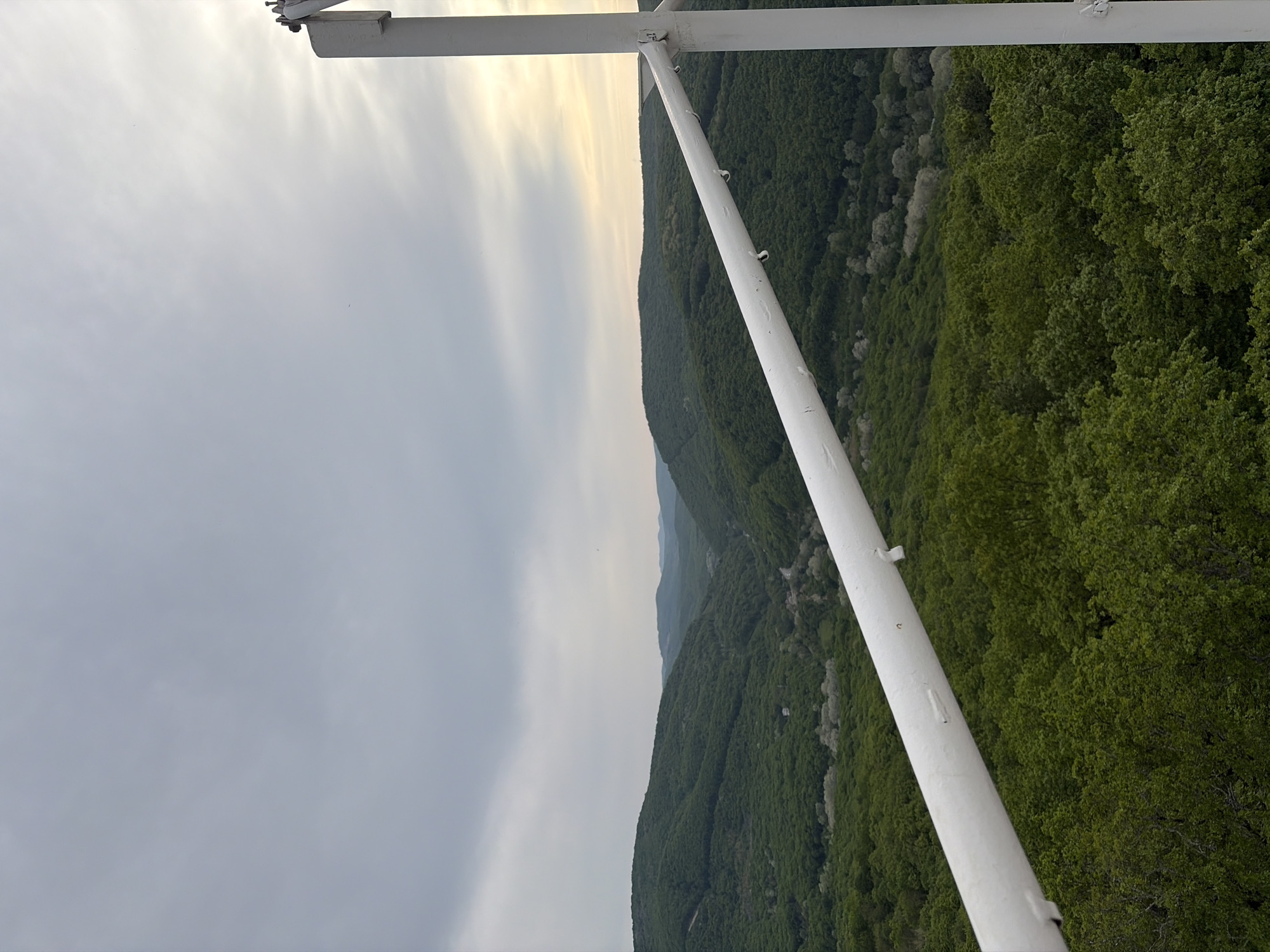 The image depicts a view from a structure, possibly a tower, overlooking a hilly, forested landscape. The scene appears to be located in or near the city of Goryachiy Klyuch, Russia, as indicated in the prompt.
A white, metal framework is prominently featured in the foreground. Beyond the framework, a series of hills are visible, covered in dense green foliage. The sky is overcast with varying shades of grey, suggesting cloudy weather. The light is soft and diffused.
There are no people visible in the image. The primary subjects are the natural landscape and the man-made structure. The main action is observation of the scenery from an elevated position.