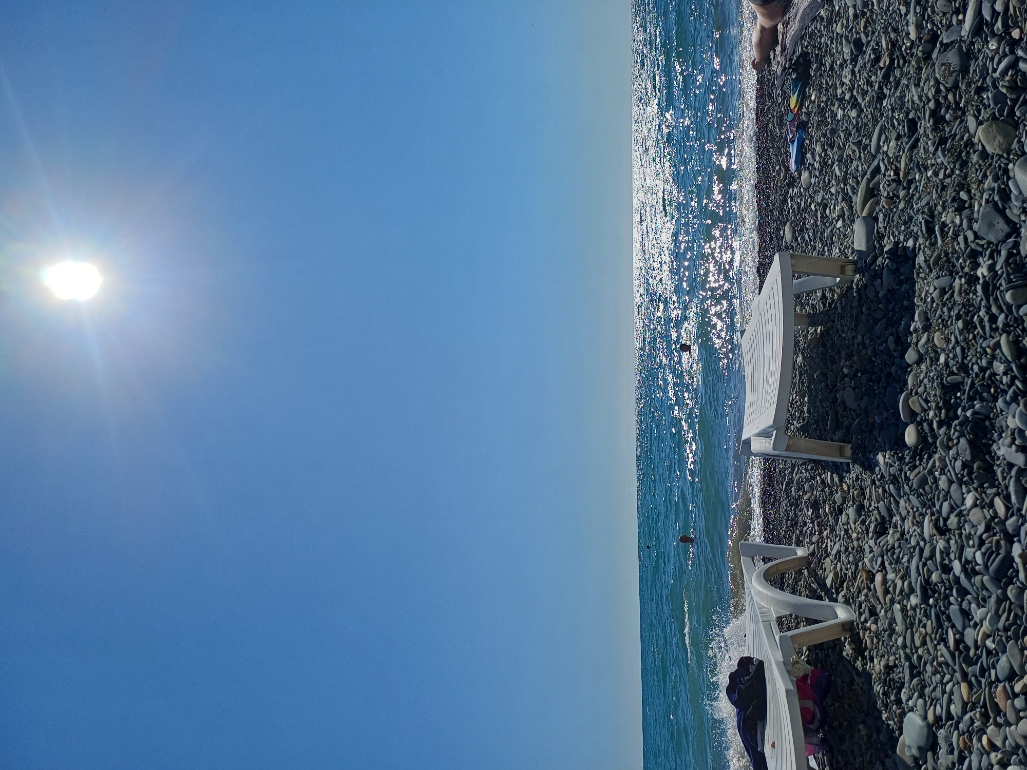 This image depicts a sunny day at a pebble beach in Sochi, Russia. In the foreground, two white plastic lounge chairs are positioned on the shore. The chair on the left has some dark clothing and a colorful bag on it. The sea, with its surface shimmering under the bright sun, stretches to the horizon under a clear blue sky. In the water, a few people can be seen swimming. The beach itself is covered in small, smooth pebbles. The sun is high in the sky, casting a bright glare.
