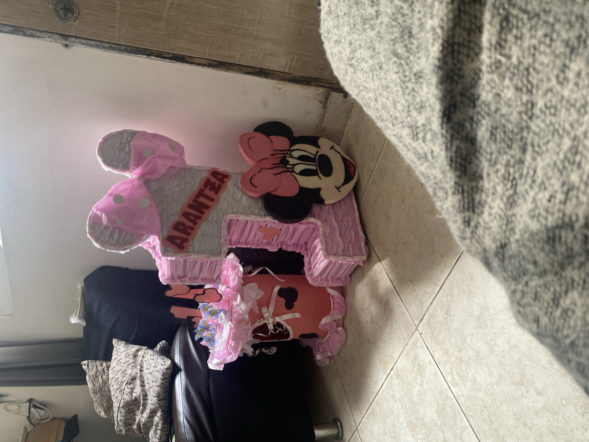 A Minnie Mouse-themed piñata with the name "ARANTZA" across the middle rests indoors on what appears to be a black leather chair with throw pillows. The floor is tiled. A gray fabric is visible in the top right corner. The location is unspecified; no evidence suggests Cotoperíz, Venezuela.
