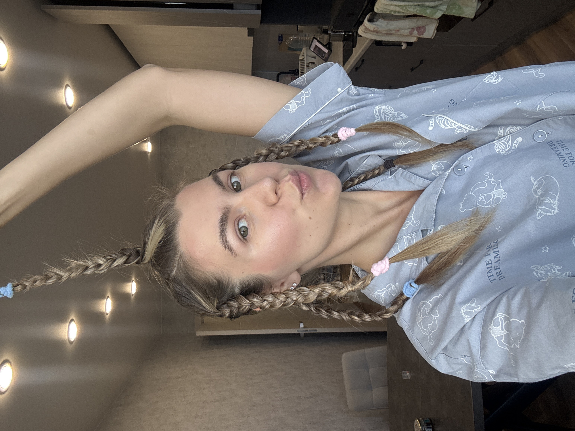 A woman in light blue pajamas with cartoon characters on them is angled in the center of the frame. Her hair is in braids with pink and blue hair ties. Her right arm is raised. The background includes a wall with embedded lights, a kitchen cabinet, and a chair. The location is indoors. There is no city or area mentioned in the image.