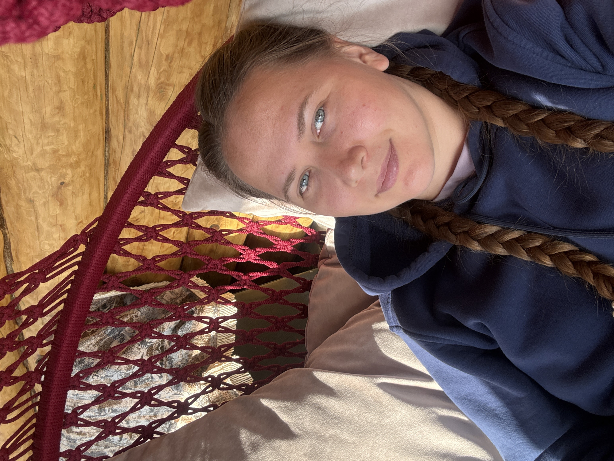 A woman with blue eyes and braided hair is lying in a maroon hammock. She is wearing a navy blue hooded sweatshirt. The hammock is suspended in a room with wooden walls. The hammock is placed above a cushion. The image appears to be taken in an indoor setting. There is no discernible information to ascertain its location as Tyulyuk, Russia.