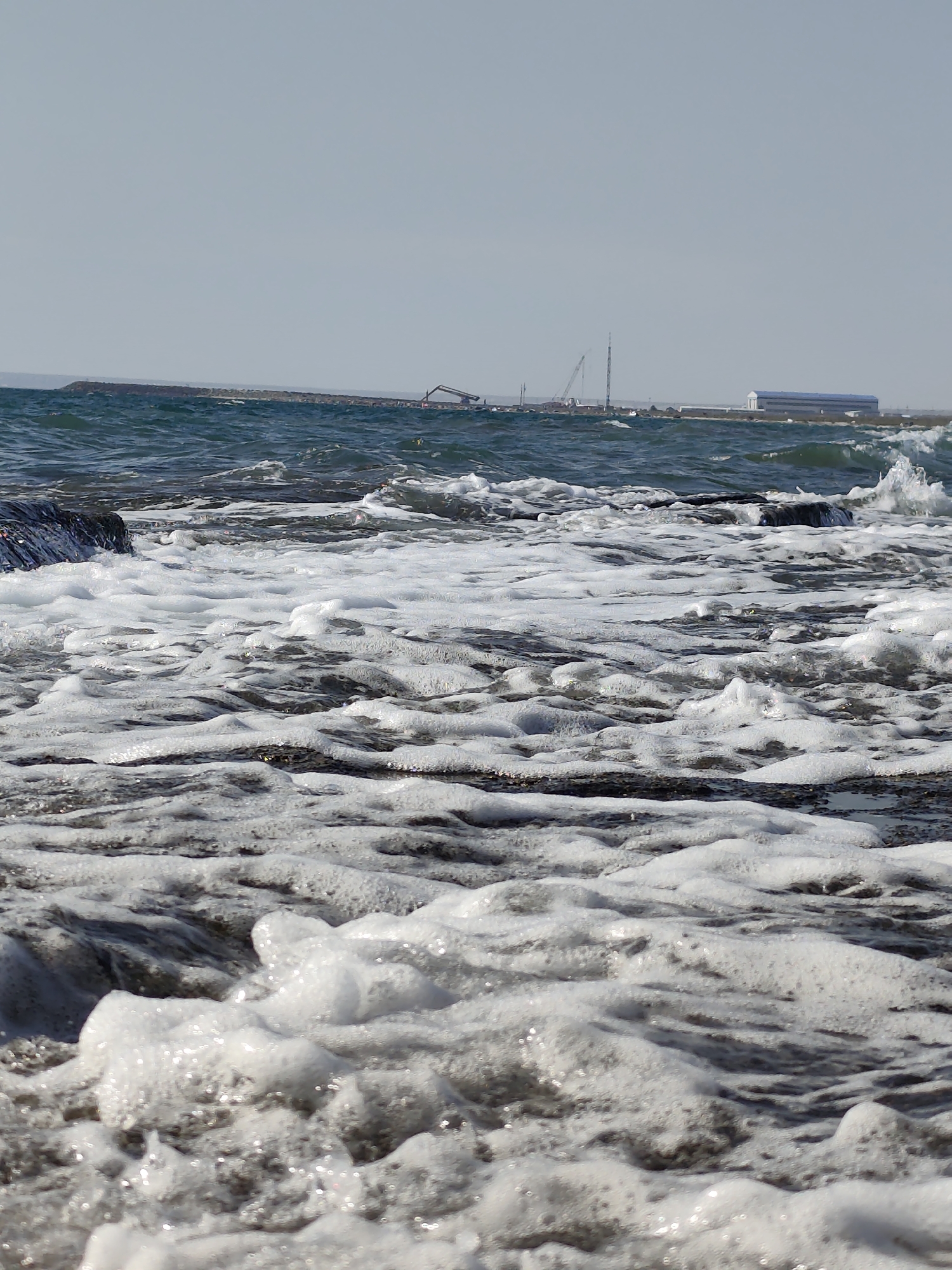 The image depicts a rocky shoreline with waves crashing onto the shore, creating white foam. In the background, there is a long breakwater with construction equipment, including cranes, visible. Further in the distance, a large building with a blue roof can be seen. The sky is clear and blue, suggesting a sunny day. The location is identified as a section of the shoreline in the city of Saiyn, Kazakhstan, likely a coastal area or a port under development. There are no people visible in the image. The scene is dominated by the natural elements of the sea and rocks, juxtaposed with the industrial presence of the construction site.