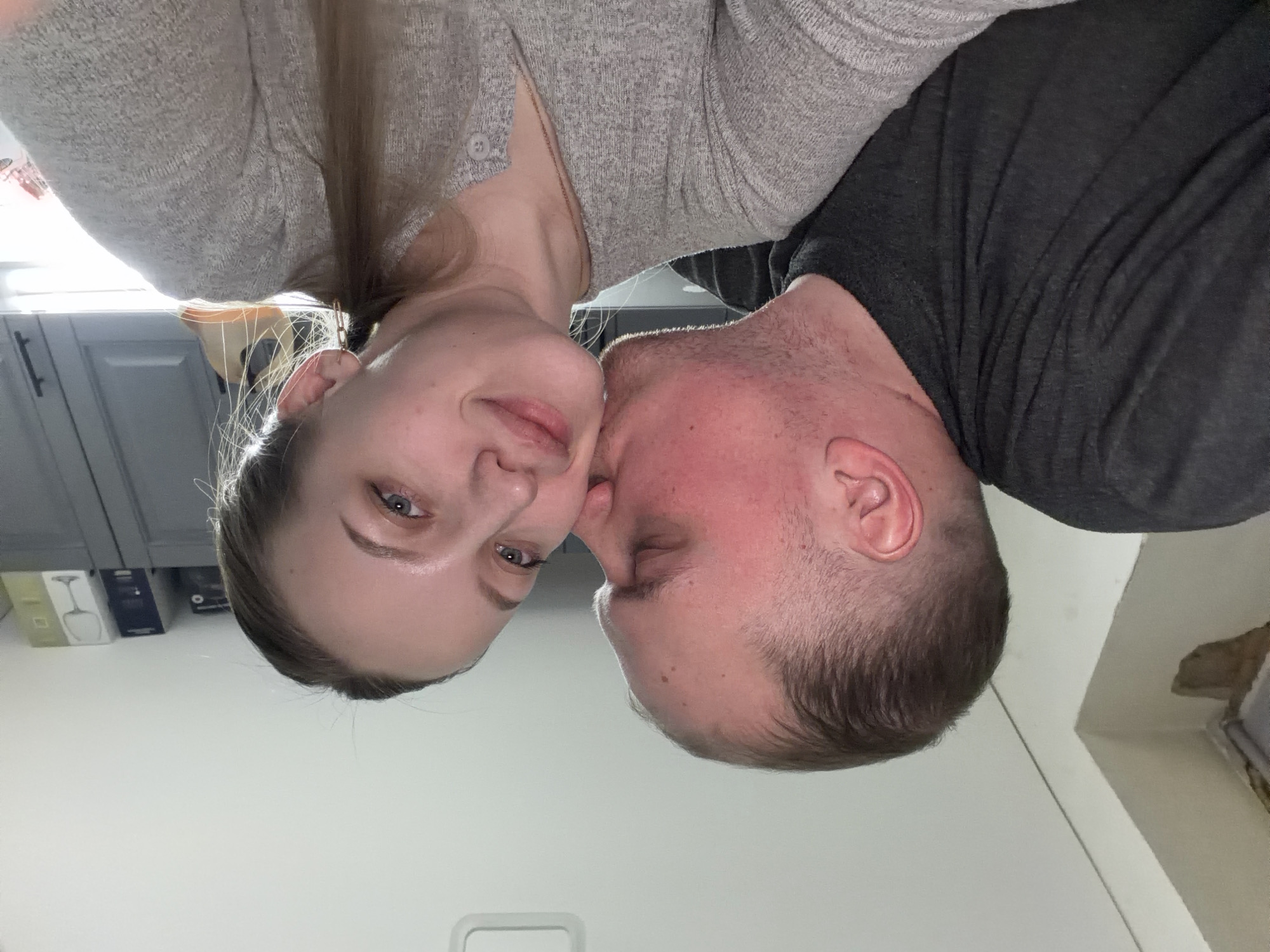 A selfie photograph depicts a light-skinned woman with light brown hair and blue eyes in a grey top nuzzling heads with a light-skinned man in a grey shirt. The background shows a kitchen with grey cabinets and boxes on shelves. The location is uncertain but the presence of boxes in the background suggest the possibility of Reshetnikovo, Russia.