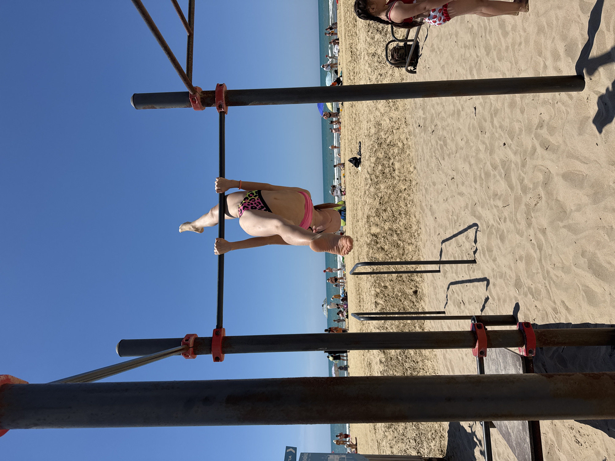 A young woman is performing an acrobatic maneuver on a pull-up bar at a beach. She is upside down, with her legs extended upwards and her hands gripping the bar. She is wearing a pink and black leopard-print bikini bottom and a pink bikini top. The background shows a sandy beach with people relaxing and the ocean. The scene appears to be a public outdoor gym setup. The image is taken from a low angle, looking up at the woman.