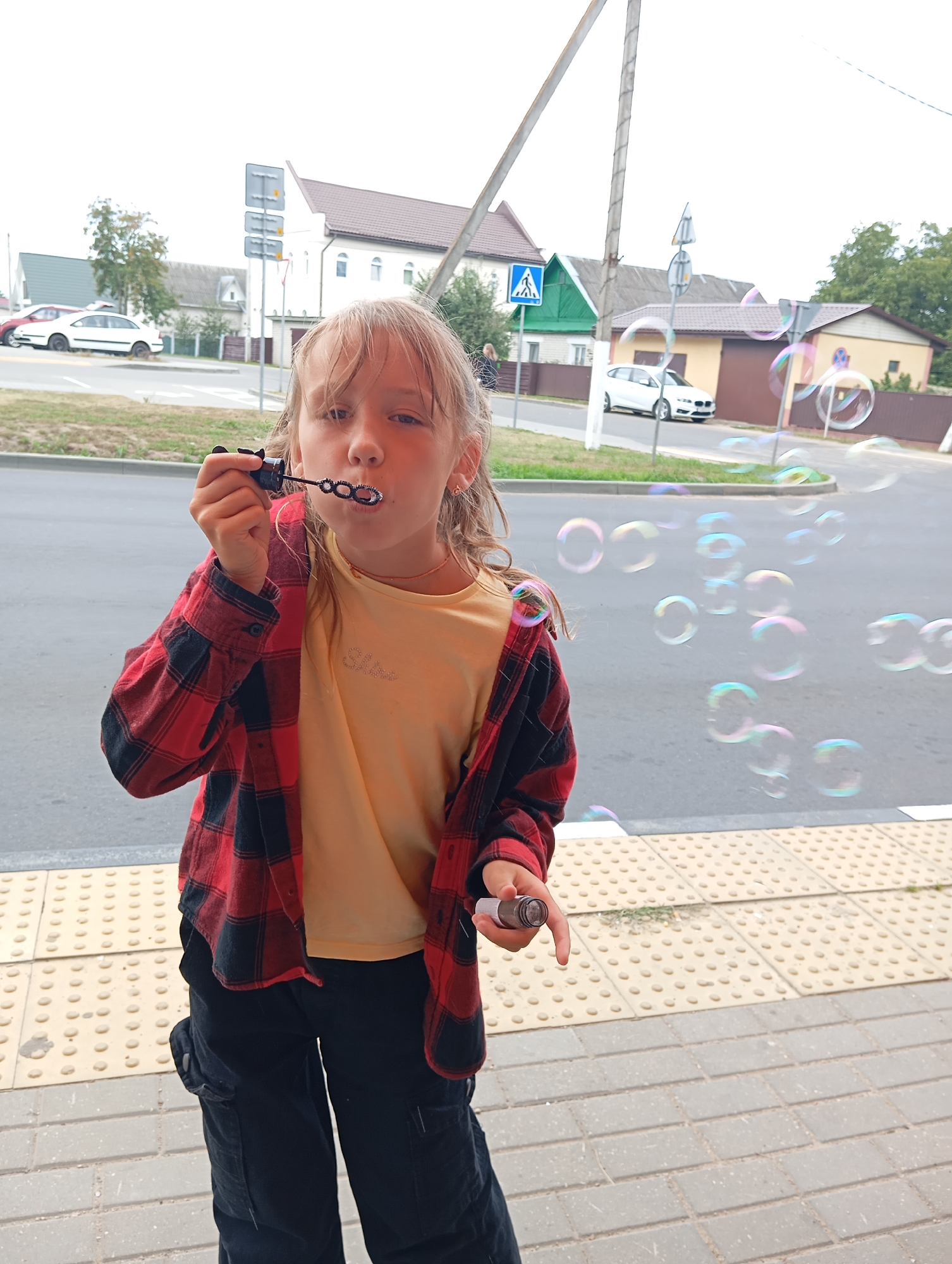 A young girl with blonde hair, wearing a red and black checkered shirt over a yellow t-shirt and black cargo pants, stands on a sidewalk and blows bubbles. She holds a bubble wand with a black handle in her right hand, and several colorful bubbles are floating in the air in front of her. The background shows a street with cars, houses, and trees. The sidewalk has a yellow tactile paving pattern.