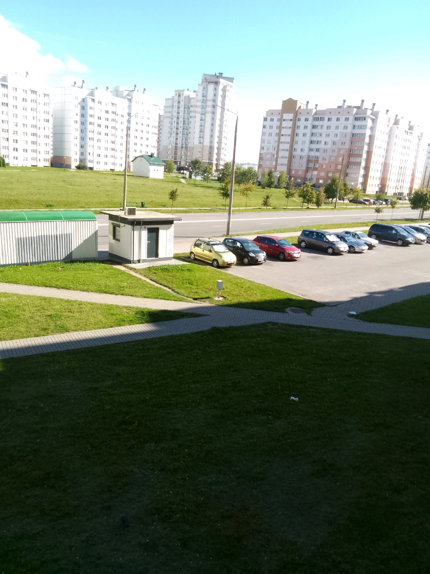 The image depicts a residential area in Hrodna, Belarus, on a sunny day. In the foreground, a well-maintained lawn is visible, bordered by a paved walkway. To the left, a long, white structure with a green roof, possibly a storage unit or garage, stands. Adjacent to it, a small, white, shed-like building with a dark door and a window is situated.
In the middle ground, a line of parked cars is visible along a paved area. These include a yellow hatchback, a dark-colored hatchback, a red hatchback, a grey hatchback, and several other vehicles. Behind the parking area, a road runs horizontally across the image, with trees and green grass lining the other side.
The background is dominated by several tall, multi-story apartment buildings, characteristic of urban residential developments. The buildings are primarily white or light-colored, with numerous windows and balconies. The sky above is clear and bright blue, with a few wispy clouds. The overall impression is one of a typical urban environment in a modern city.