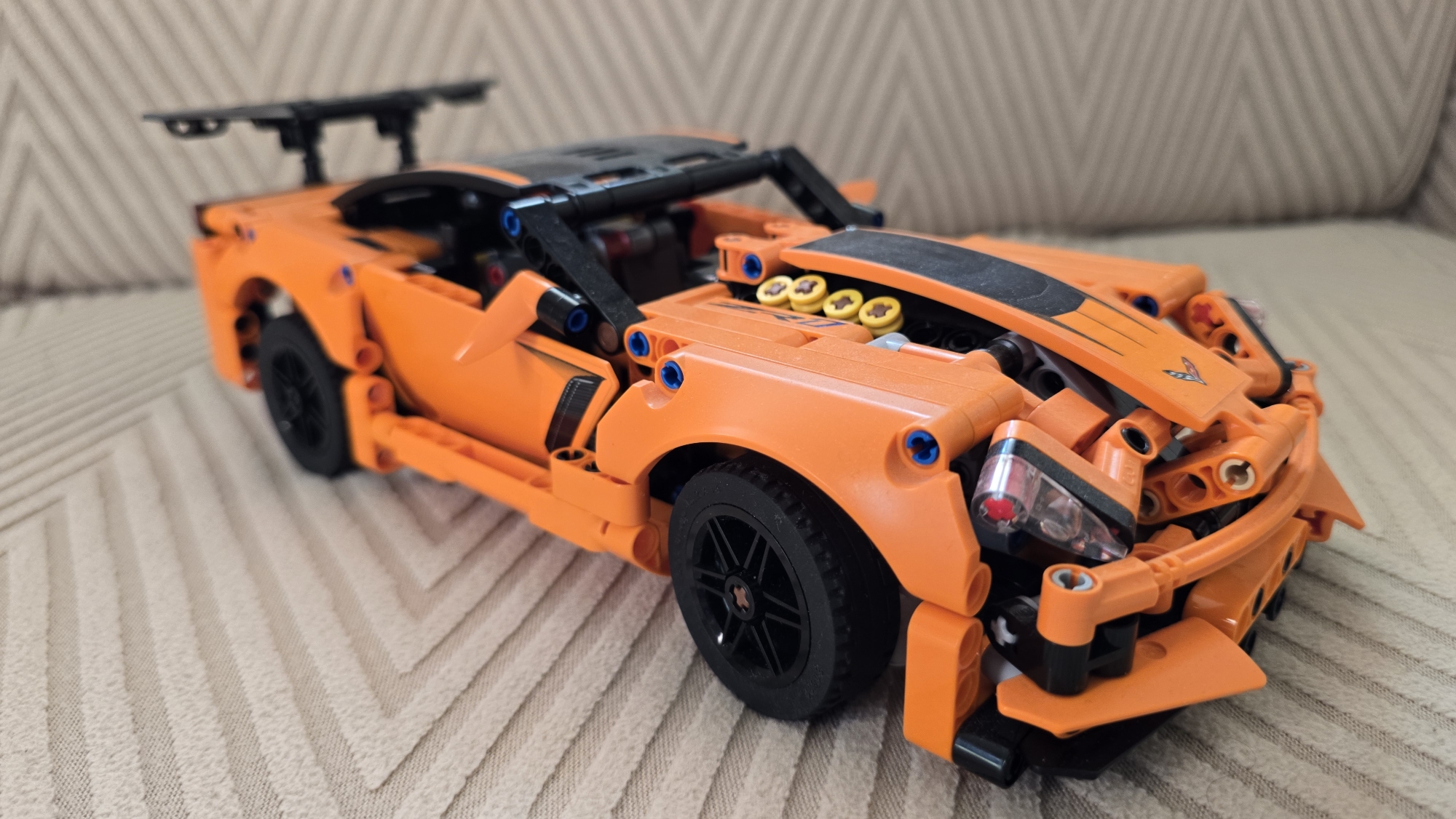 A Lego Technic Chevrolet Corvette C8 Stingray, predominantly orange with black accents, is positioned on a beige, textured fabric surface, likely a sofa or chair. The toy car is oriented diagonally, with its front-left side closest to the viewer. The car features visible building block construction, including exposed Technic pins and gears. The background consists of the same beige fabric. There is no indication of the city of Верхнеказымский, Russia, or any other specific location within the image.