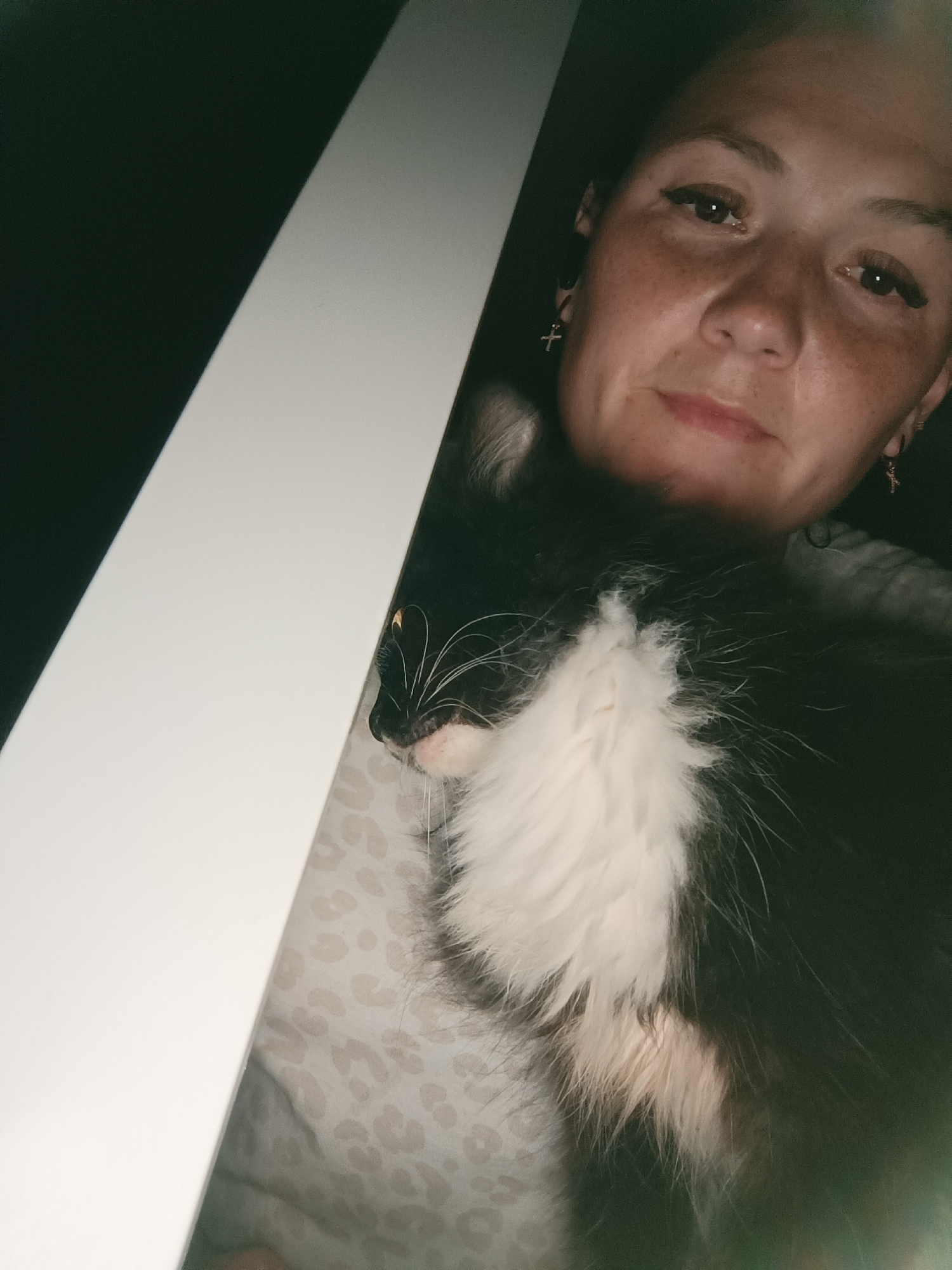 In this close-up, low-light, indoor selfie, a woman with light skin, freckles, and blonde hair pulled back is holding a black and white cat. The woman is looking towards the camera with her eyes open, wearing makeup including eyeliner and mascara. She has small cross-shaped earrings in both ears. The cat is nestled against her chest, with its head turned slightly to the side, revealing its whiskers and a patch of white fur on its chest. The background is dark, with a diagonal white object on the left side of the frame. The scene is likely within a residential setting in Chaikovsky, Russia.