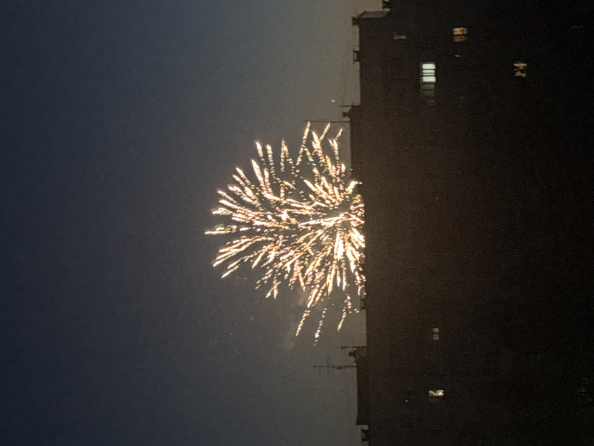 A single, large burst of yellow and white fireworks is centered in a dusky blue sky, appearing like a radiant dandelion. The fireworks are positioned above a dark, silhouetted cityscape, suggesting a nighttime urban event. The lower portion of the image is dominated by the rooftops and facades of apartment buildings, some with faintly illuminated windows. A tall antenna structure is visible on the left side of the buildings. The overall scene captures a moment of celebration or spectacle against the backdrop of a residential area in Chaikovsky, Russia.
