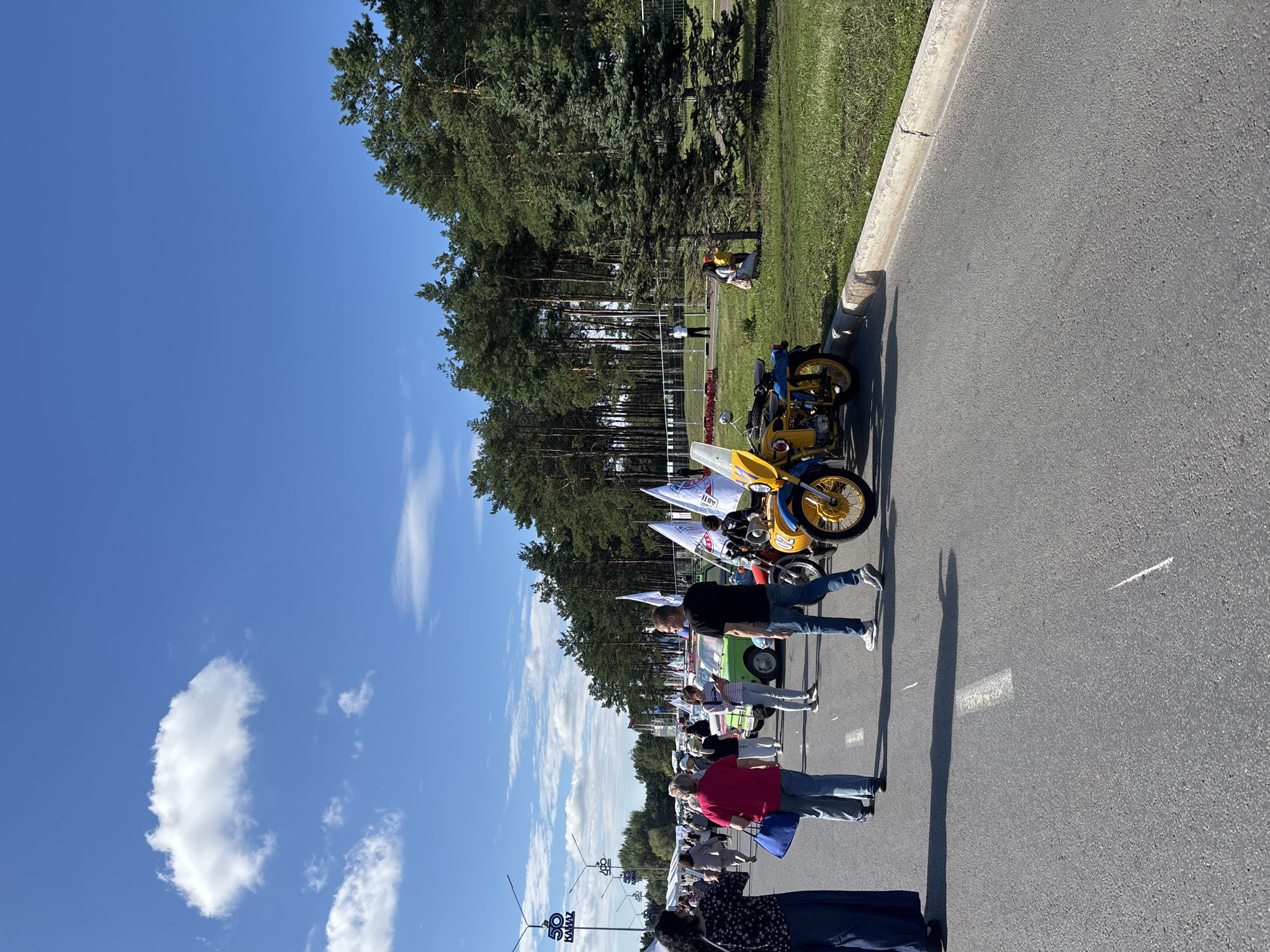 A sunny outdoor event, possibly a car show or festival, is depicted in Naberezhnye Chelny, Russia. The scene features a paved road with a white lane marker, lined by a grassy area and a dense forest of tall pine trees. Several people are walking on the road, some appearing to be spectators and others possibly participants.
In the foreground, a yellow and blue motorcycle with a sidecar is parked, displaying a license plate with the number "02". Further down the road, a light green vintage car is visible. Numerous flags with white fabric are displayed on poles, some appearing to be promotional banners. In the background, there are tents or stalls set up. Overhead, a cable car system with blue signage is present. The sky is bright blue with scattered white clouds. The overall impression is of a public gathering in a park-like setting, possibly related to the automotive industry given the "KAMAZ" signage visible on a blue pole.