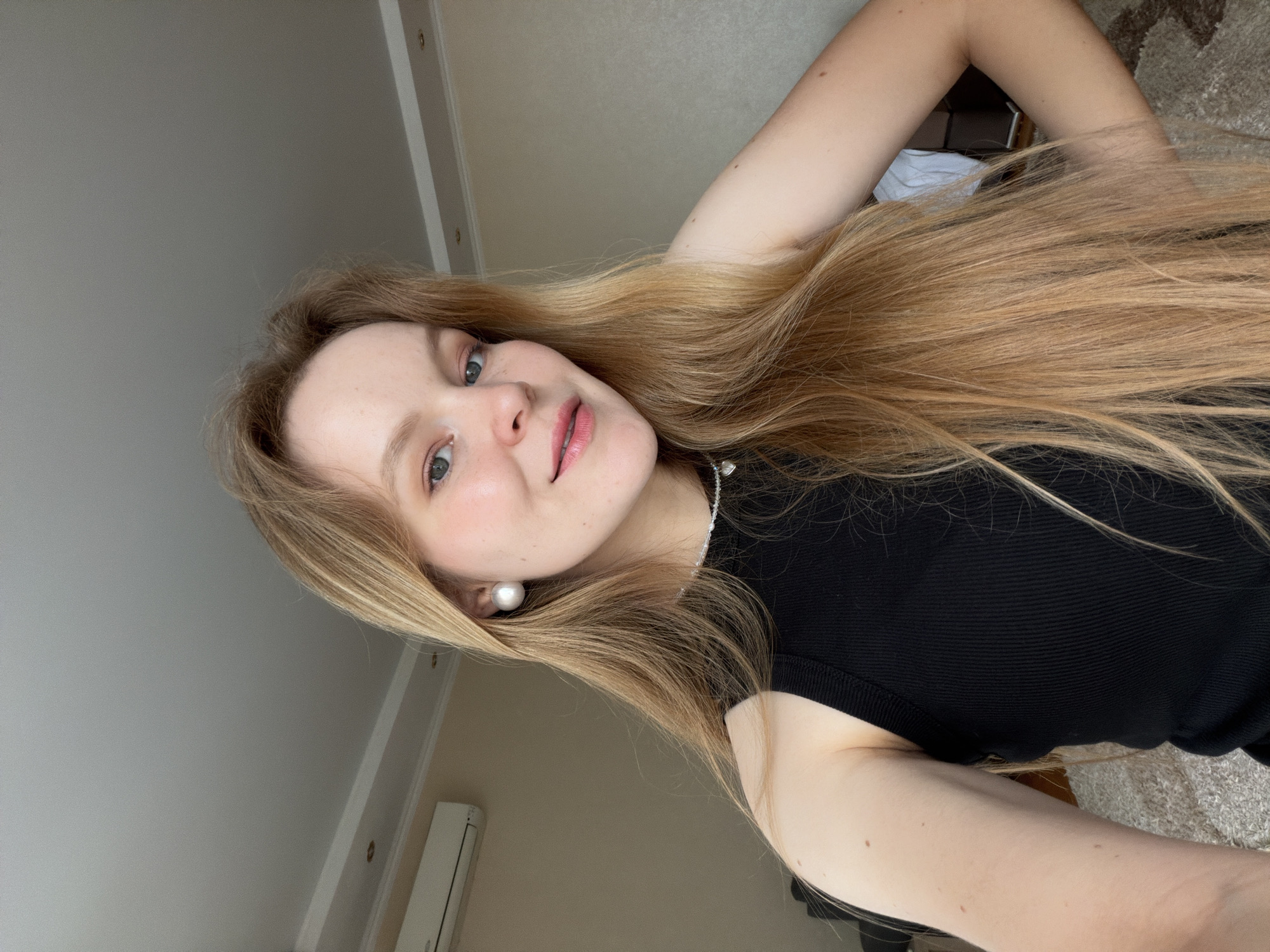 A young woman with long blonde hair and blue eyes is pictured indoors. She is wearing a black sleeveless top and large pearl earrings. Her left arm is bent at the elbow with her hand on her hip, and her right arm is out of frame. She is looking at the camera with a slight smile. The background shows a wall with a air conditioning unit and a ceiling with ornate molding. The floor is covered with a textured rug. The image appears to be a selfie taken in a residential setting. There is no information to place this image in Kazan, Russia.