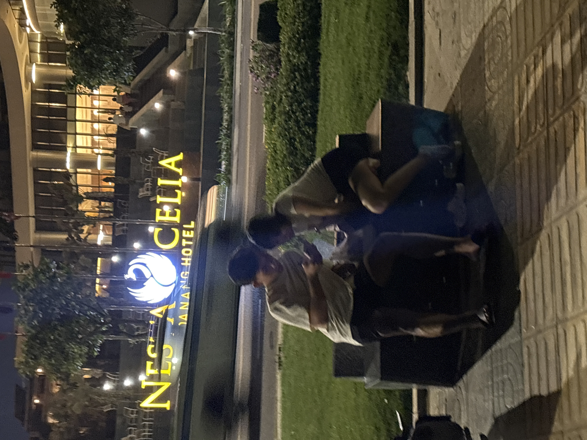 Two people, possibly adults, are huddled around a dark object on a sidewalk next to a patch of green grass in front of the Nesta Hotel in Đà Nẵng, Vietnam. The sidewalk is tiled. One person is wearing a light-colored t-shirt and dark shorts, while the other is wearing a darker t-shirt and dark shorts. The object they are focused on is dark and indistinct. In the background is a sign that reads "NESTA CELIA DANANG HOTEL" in yellow letters with a swan logo. The hotel is multi-storied and illuminated. The environment appears to be at dusk or nighttime.