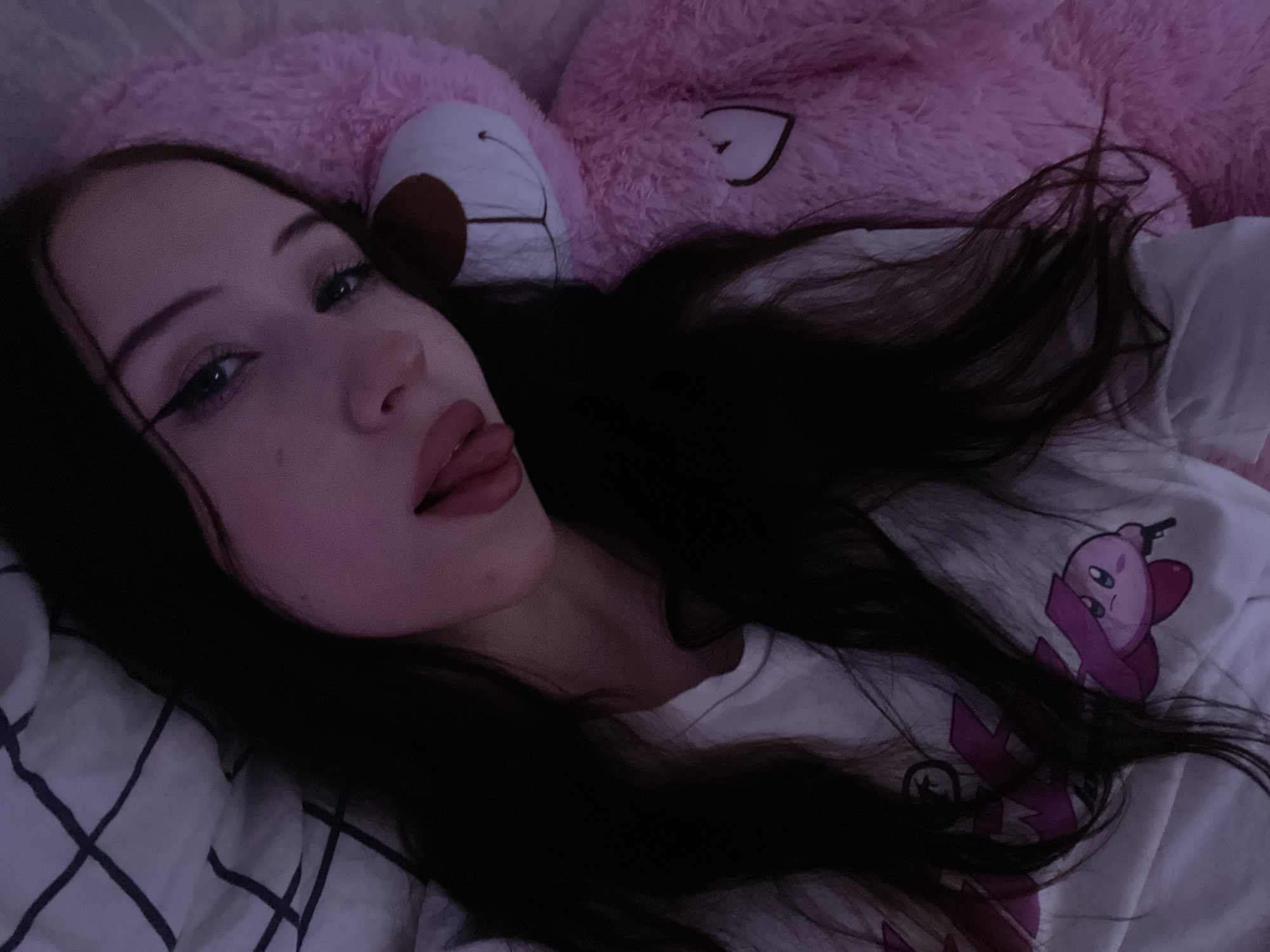 In the city of Votkinsk, Russia, an image captures a close-up of a young woman with long, dark hair lying down. She is wearing a white t-shirt with a pink character graphic, possibly Kirby, on it. She has winged eyeliner and her tongue is sticking out slightly. To her right is a large, fluffy pink teddy bear, which appears to be a significant portion of the frame. The background shows a checkered pattern, suggesting she is in a bed with patterned sheets. The lighting is dim, with a purple-pink hue, contributing to a soft and intimate atmosphere.