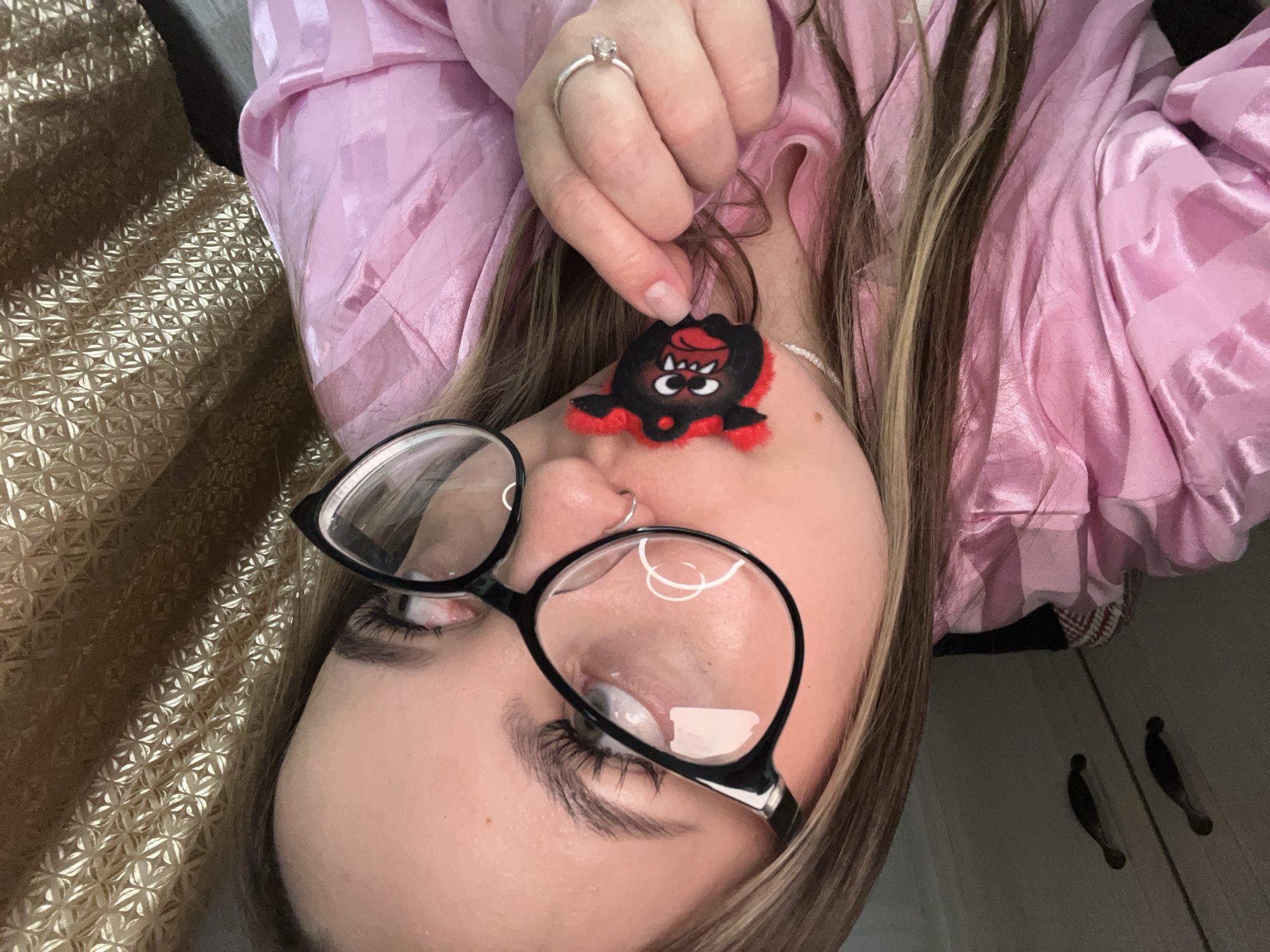 A person wearing glasses and a pink satin robe is holding a small, black and red fuzzy monster keychain in their hand. The person has brown hair with blonde highlights, a nose ring, and a ring on their finger. The background includes a gold textured surface and a white door with dark handles. The location is not discernible from the image.