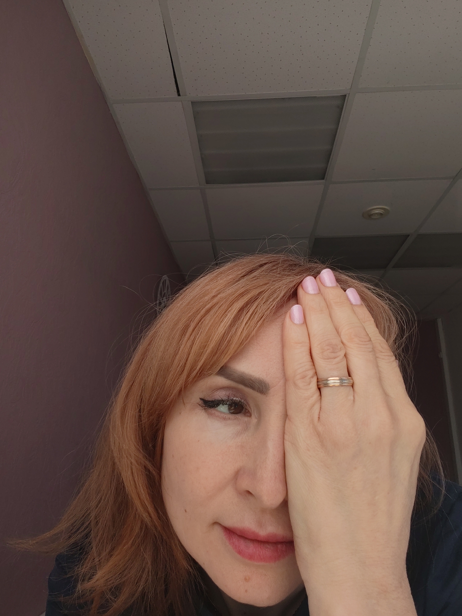 The photograph shows a woman indoors, with her right hand covering her right eye. She has reddish-brown hair, fair skin, and is wearing a ring on her right ring finger. The visible portion of her face shows an eye with eyeliner and lipstick. The background consists of a pink wall and a drop ceiling with fluorescent lighting. The location is not discernible as Chaikovsky, Russia, from the provided image alone.