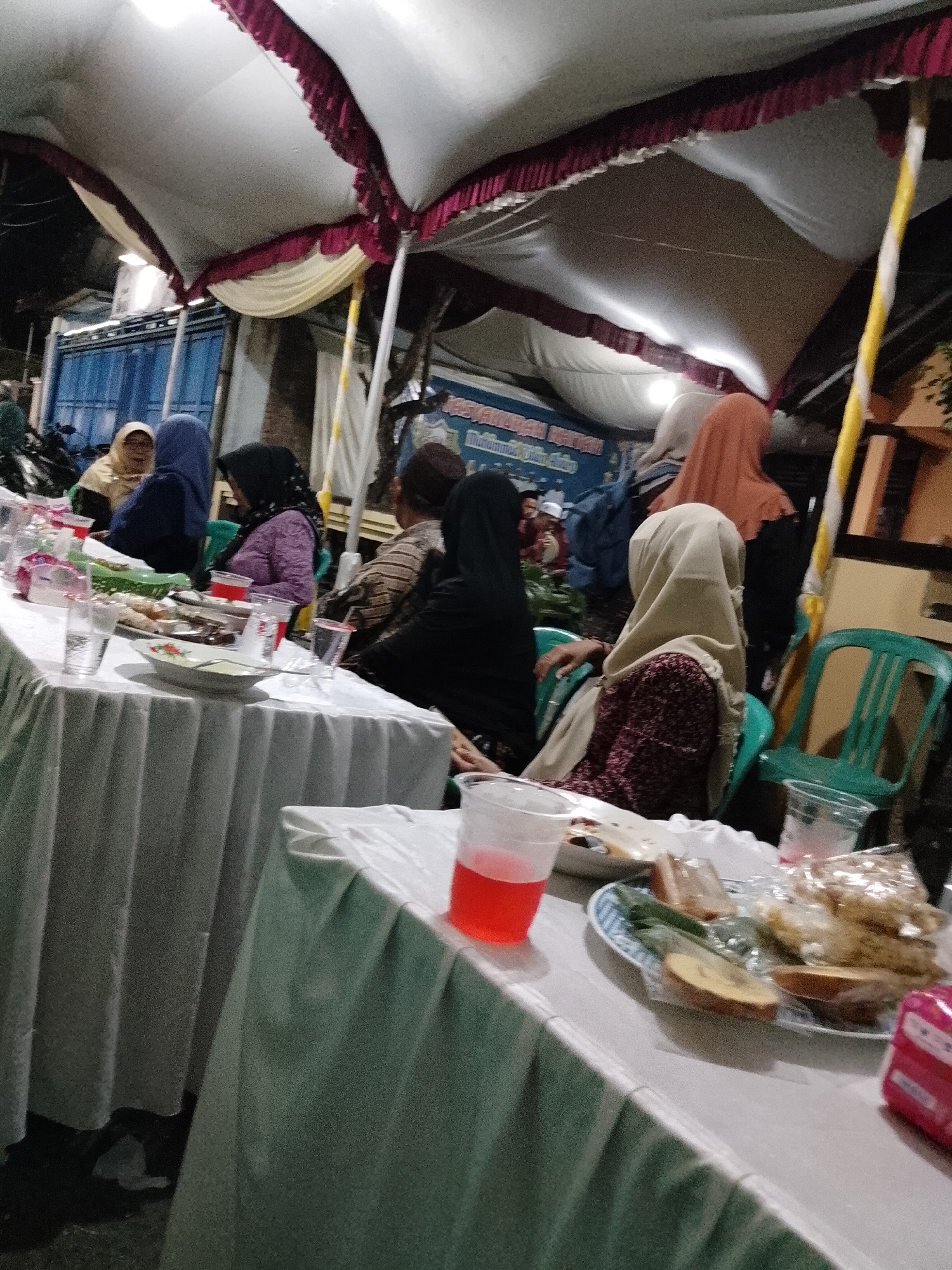 The image depicts an outdoor event, likely a meal or gathering, held under a white tent with maroon and yellow trim. Several individuals, predominantly women wearing hijabs, are seated at tables covered with white tablecloths. The tables are set with plates of food, glasses, and beverages, including a red drink in a clear plastic cup prominently visible in the foreground. In the background, a blue corrugated metal structure and some vegetation can be observed. The scene appears to be at night, with artificial lighting illuminating the area. The presence of a blue and white banner with text, partially visible in the background, suggests a communal event, possibly a wedding or a religious celebration. While the specific location within Trangkil, Indonesia, is not definitively identifiable from the image alone, the overall context suggests a local community gathering.