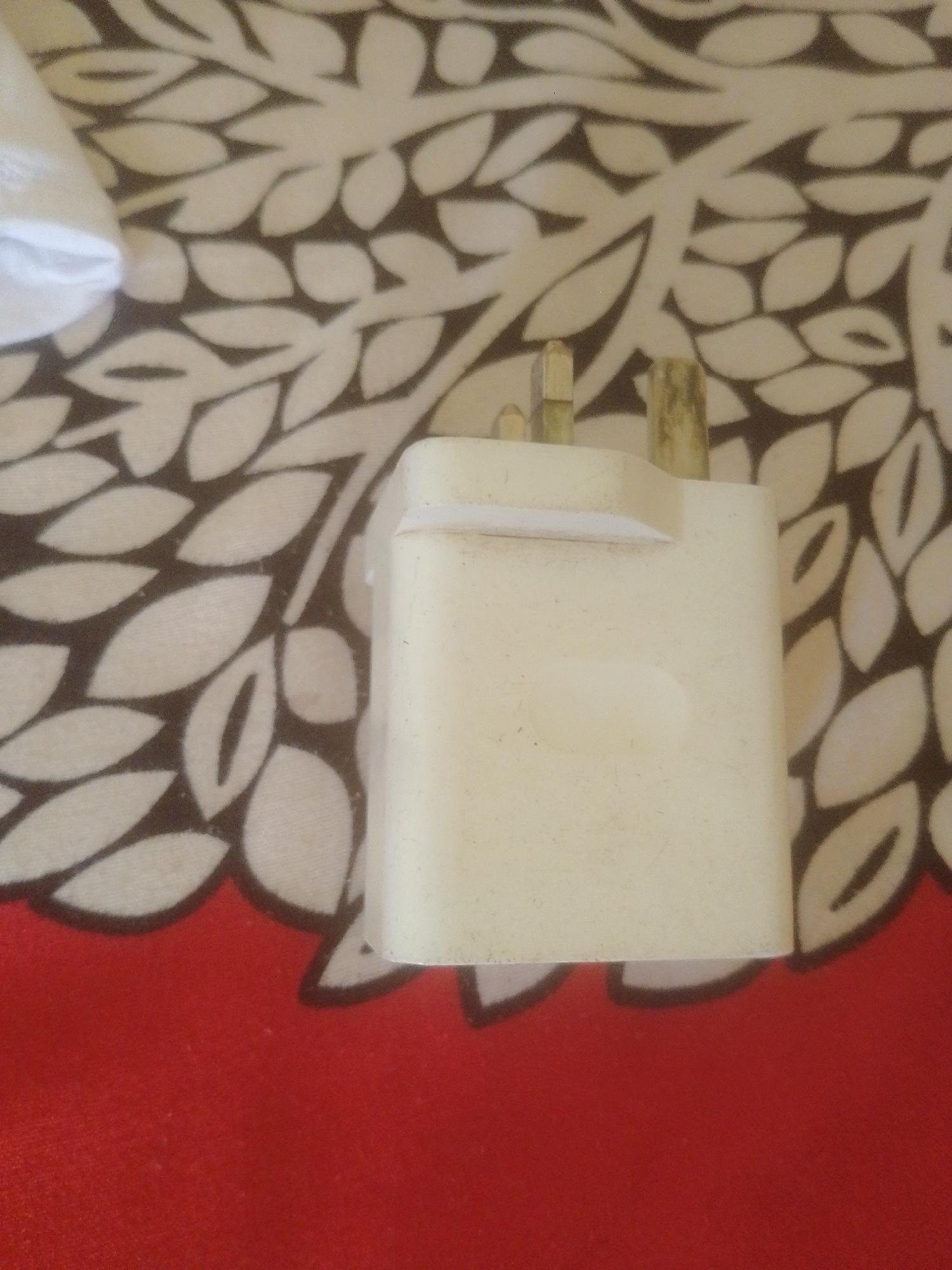 A close-up shot shows a white electrical plug resting on a patterned fabric surface. The fabric features a repeating design of white leaf-like shapes against a dark background, with a section of solid red fabric at the bottom of the image. The plug itself is white and appears to be a universal adapter, with three prongs visible. The lighting is somewhat dim, casting soft shadows. The location in Danbazau, Nigeria, is not discernible from the visual information provided in the image.