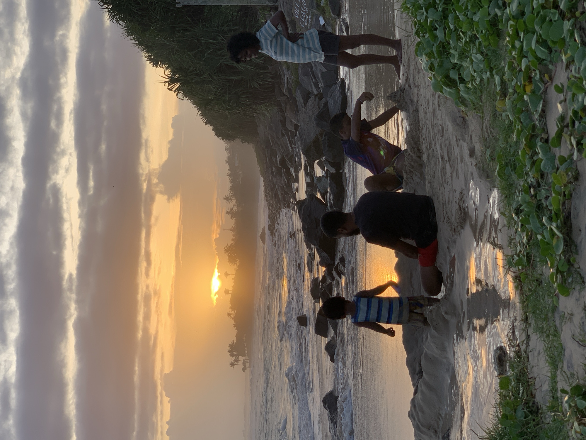 This image depicts a coastal beach scene at sunset in Kathaluwa, Sri Lanka. The sun, a bright golden-orange orb, is low on the horizon, partially obscured by distant landforms and low clouds, casting a strong reflection across the ocean and wet sand. The sky above features layers of pale orange and grey clouds.
In the foreground, four individuals are present on the sandy shore. On the far left, a young child, viewed from the back, wears a blue and white striped shirt and light-colored shorts; they stand in shallow water near a sand mound. To their right, an adult male sits on the sand, also facing away from the viewer, wearing a dark t-shirt and red shorts, appearing to interact with the sand mound. A second child, positioned to the right of the seated male, is crouched on the sand, actively shaping a smaller sand structure with their hands. This child wears a dark t-shirt with a colorful design. On the far right, an adult female stands upright, facing left towards the group, with her right hand on her hip. She wears a light-colored horizontally striped t-shirt and dark shorts, and is barefoot.
The shoreline is characterized by numerous dark, irregular rocks extending into the breaking waves of the ocean. The water exhibits a golden sheen from the sunset. Along the right side of the frame, dense, dark green coastal vegetation, likely pandanus or similar shrubs, grows upwards from the beach. Scattered green ground cover with round leaves is also visible on the sand in the immediate foreground. In the distant left horizon, silhouetted palm trees and faint outlines of structures are visible, with a small building or gazebo identifiable amidst the trees directly beneath the setting sun. A partially visible white sign with text, possibly "VILLA CLOUD," is located behind the standing female near the vegetation line.