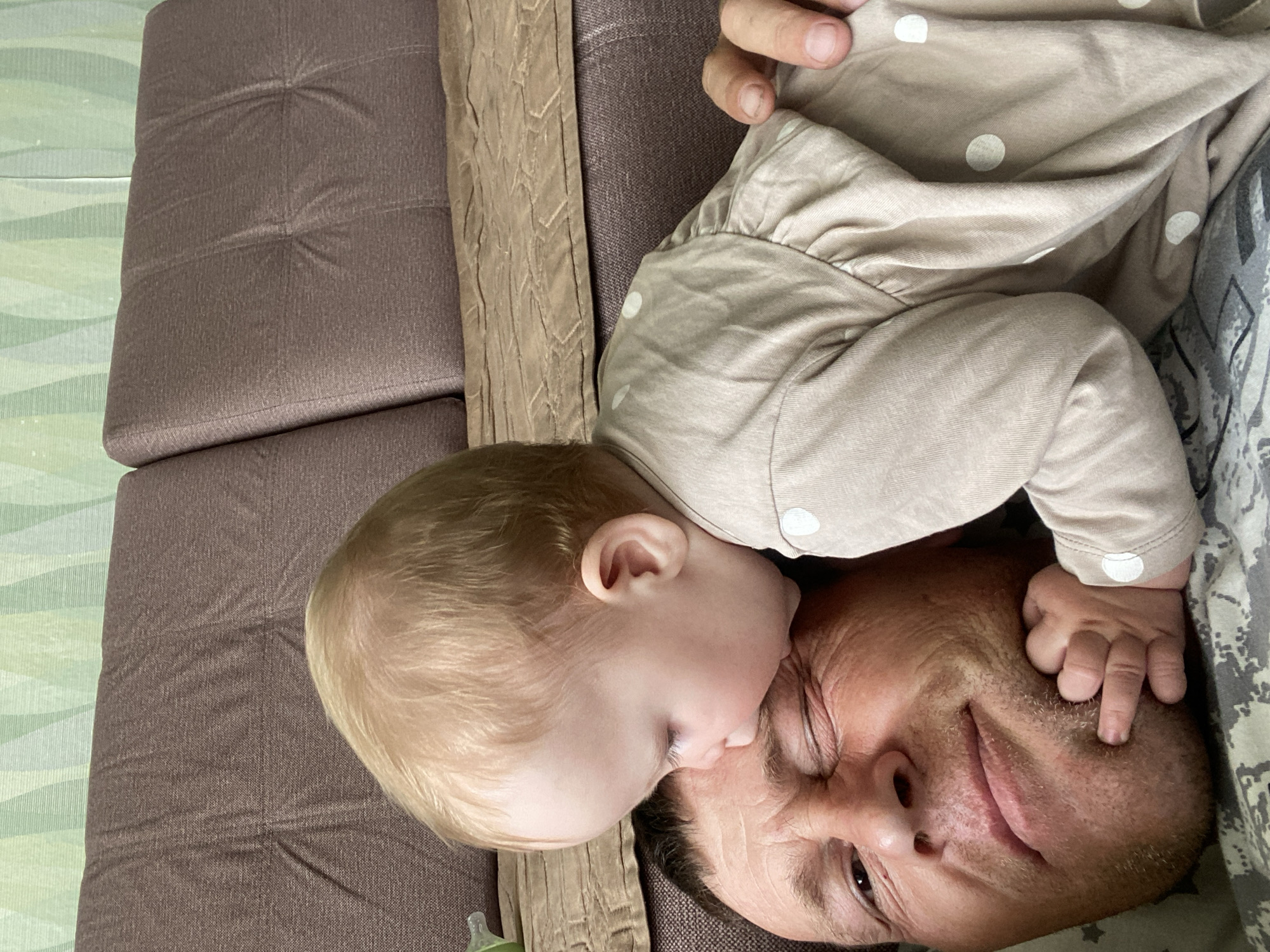 A close-up, overhead shot shows a baby's face nuzzling into the forehead of a man who is lying on his back. The man's eyes are closed or squinting, and he has stubble on his face. The baby, with blonde hair, is wearing a light-colored onesie with white polka dots. The man is lying on a patterned surface that appears to be a bed or sofa. In the background, a portion of a brown sofa is visible, with a green and white patterned wallpaper above it. The scene appears to be indoors, within a home environment in the city of Fokino, Russia. The interaction suggests a moment of affection between the baby and the man.