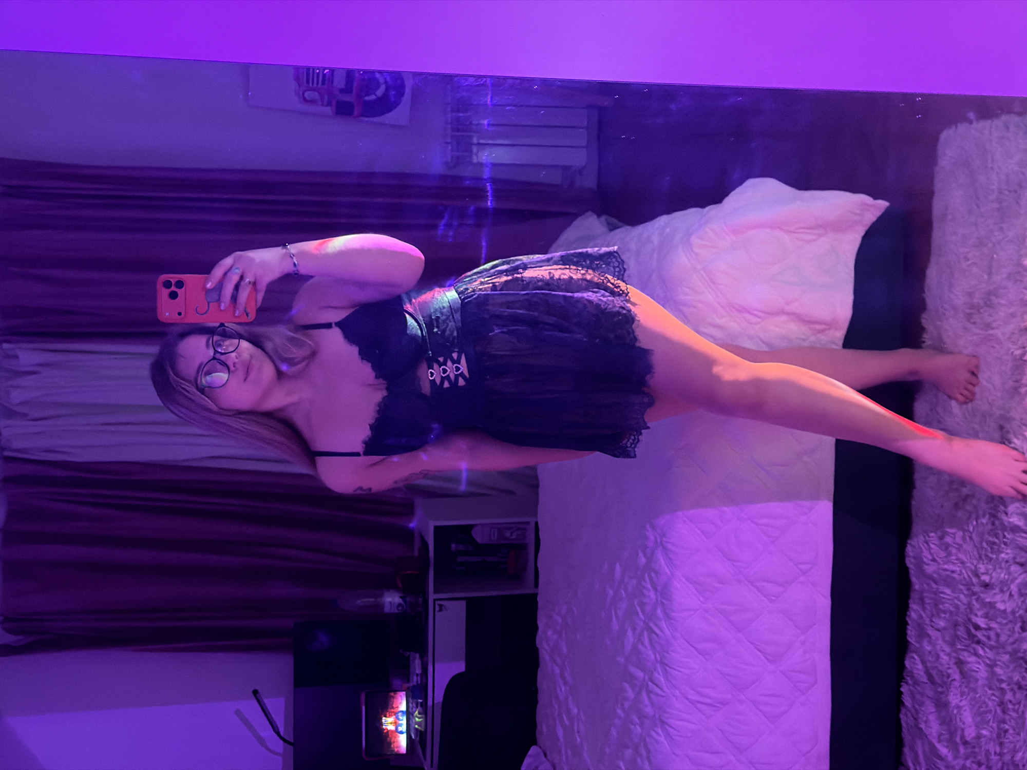 The image depicts a woman in a bedroom in Большая Шильна, Russia, taking a selfie in a mirror. She is wearing glasses and a black lace dress with a black belt. She is holding a red cell phone. The room has a bed with a white comforter and a purple curtain. There is also a white shelf with various items on it and a computer screen. The room is lit with purple light.