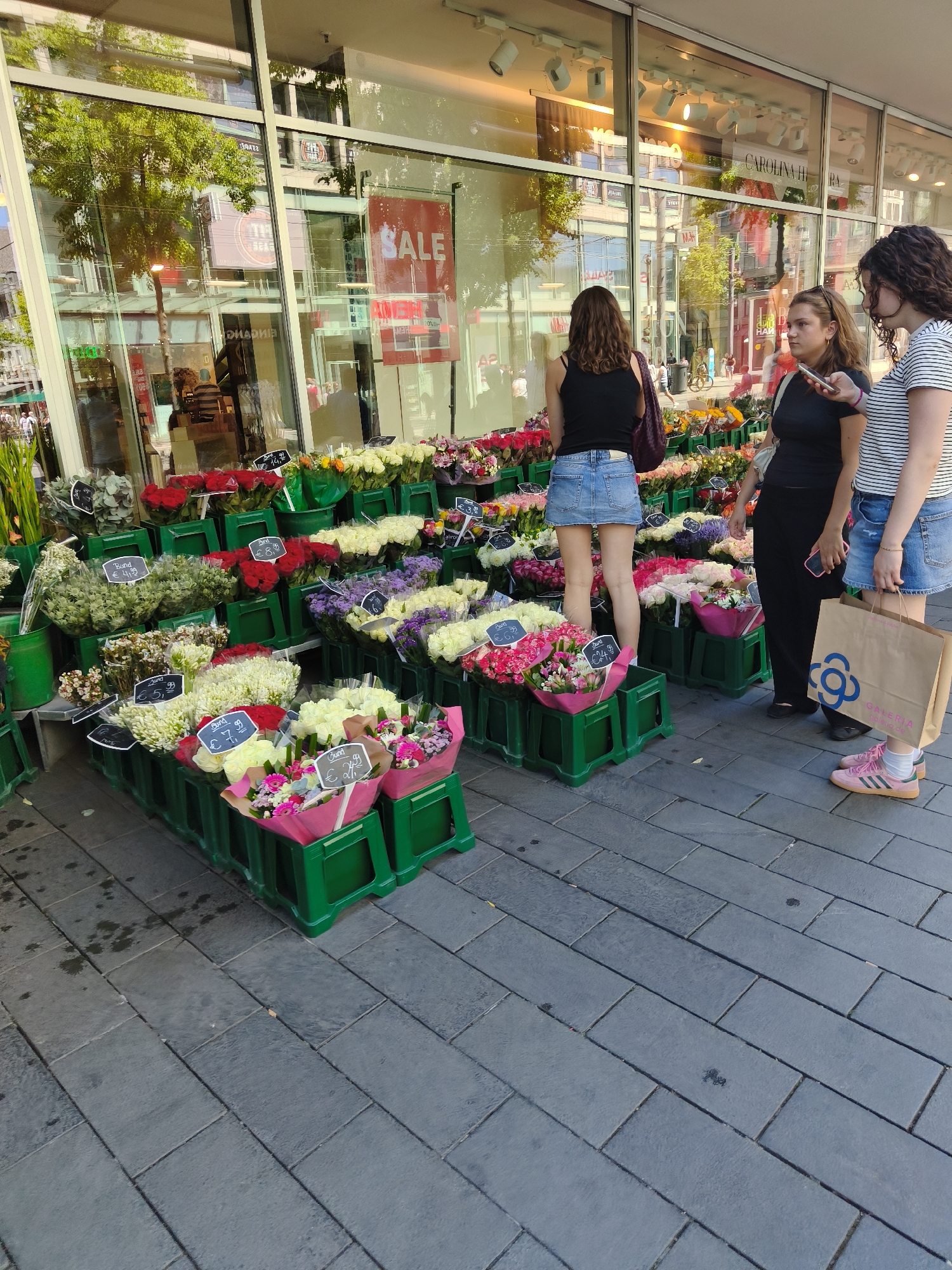 Three young women stand on a stone-paved pedestrian area in front of a store with large glass windows. The store appears to be a clothing boutique, with mannequins and clothing displays visible through the windows. One woman on the left wears a black tank top and a denim skirt, looking at a display of flowers. The middle woman, wearing a black t-shirt and black pants, holds a smartphone and looks at it. The woman on the right wears a striped t-shirt and denim shorts, holding a shopping bag from "GALERIA".
In front of the store is a vibrant display of cut flowers arranged in green plastic crates. There are various types and colors of flowers, including red roses, white roses, pink roses, and purple flowers. Some bouquets have price tags attached, indicating prices like € 3.99, € 6.99, and € 7.99. The ground is paved with large rectangular dark grey stones, with some scattered water spots. Reflections of trees and buildings are visible in the store's windows, along with a red "SALE" sign. The overall scene suggests a busy shopping street on a sunny day in Mannheim, Germany.