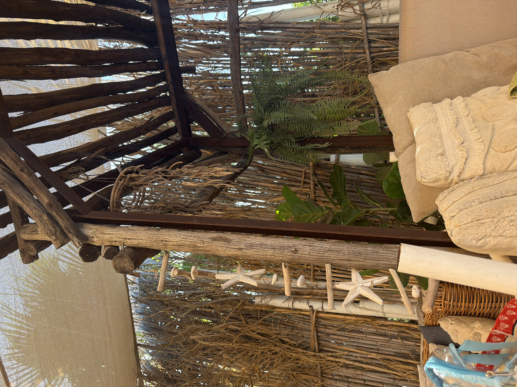 The image depicts an outdoor seating area, possibly a covered patio or gazebo, with a rustic and natural aesthetic. The structure appears to be made of weathered wood and bamboo or reeds, creating a beachy or bohemian atmosphere. A light-colored fabric canopy stretches overhead, filtering the sunlight.
Several decorative elements are visible. A hanging mobile, crafted from driftwood pieces and possibly shells or stones, is suspended from the wooden structure. Starfish motifs are incorporated into the decor, with a white starfish prominently displayed on the mobile and another partially visible lower down. Green foliage, likely a hanging fern or similar plant, adds a touch of life to the scene.
In the foreground, a plush sofa or seating arrangement is laden with various cushions. The cushions are textured, with some featuring fringe detailing and intricate embroidery in neutral tones. A woven basket sits near the sofa, and a partially visible clear plastic bag with blue straps is also present, suggesting personal belongings. The background is filled with a dense arrangement of vertical reeds or bamboo stalks, which serve as a wall or partition. The overall impression is one of a relaxed, outdoor lounging space designed with natural materials and coastal-inspired decor.
While the image strongly suggests a location with a Mediterranean or coastal climate, specific details placing it in Protaras, Cyprus, are not inherently present within the visual information provided.