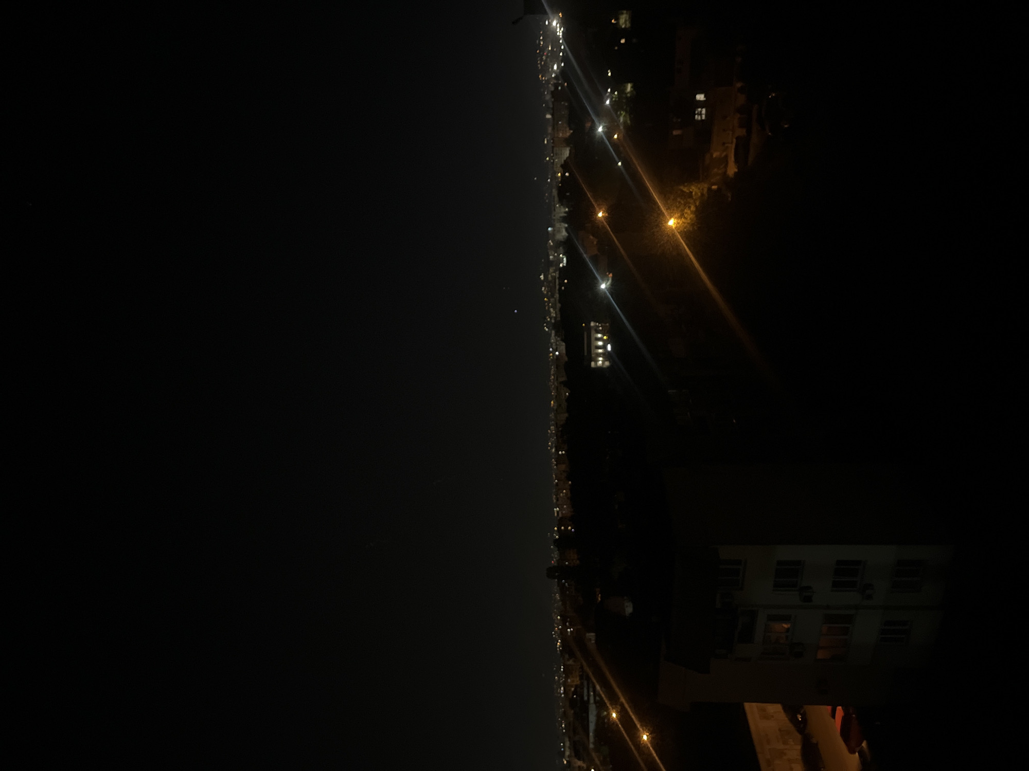 The image shows a nighttime view of a cityscape, potentially along a coastline or waterway, as evidenced by the dark expanse to the left. Along the center and right side of the image, numerous lights indicate a city or urban area. There are streetlights and building lights visible, forming a bright line against the darkness. In the foreground, the top of a light-colored building or structure is visible with illuminated windows, and cars appear to be parked nearby. The image is taken from an elevated perspective, potentially from a high-rise building or hill. The location is Sofia, Bulgaria, which isn’t on the coastline, but the image is in a rotated position and could possibly be a view of a lake or reservoir from an elevated position near the city.