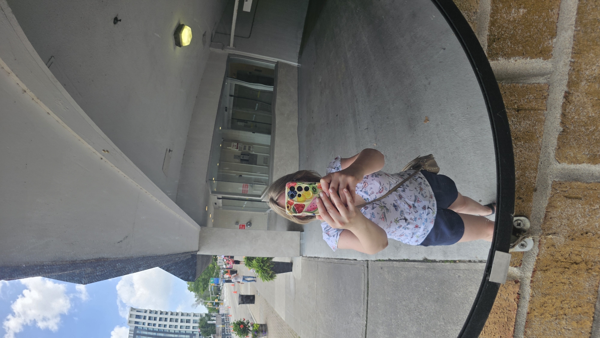 The image is a reflection in a large, round mirror. In the reflection, a person is taking a selfie with a smartphone. The person has blonde hair and is wearing a white top with a pattern of colorful drawings and dark blue shorts. A light brown purse is slung over their shoulder. The mirror reflects a building entrance with glass doors and windows, a concrete sidewalk, and a brick wall. In the background of the reflection, people can be seen walking on the sidewalk, and there are some trees and streetlights. Further in the distance, a modern white building with many windows is visible against a bright blue sky with white clouds, suggesting it is daytime and the weather is fair. The overall setting appears to be an urban or suburban environment, possibly a commercial or institutional building entrance. The reflection is distorted by the curved surface of the mirror. The image has a vertical orientation, and the content within the mirror is also oriented as if the camera were held upright. There is no visible text in the image.