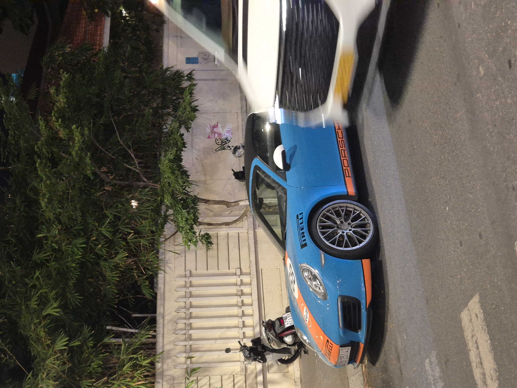 A blue Porsche 911 GT3 RS with orange racing stripes is parked on a city street at night in Đà Nẵng, Vietnam. The car is adorned with "Pirelli" and "Gulf" branding. A white van is partially visible in the foreground, obscuring part of the Porsche. In the background, a white fence separates the street from a building with bamboo trees and other foliage. A black scooter is parked near the fence, behind the Porsche. The street surface is asphalt with painted white lines indicating a pedestrian crossing. The lighting suggests it is nighttime, with artificial lights illuminating the scene.