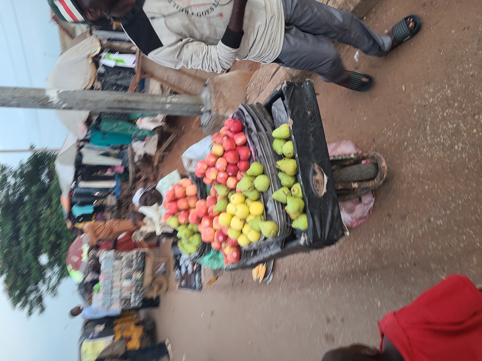 In the city of Nyanya, Nigeria, a street scene unfolds with a focus on a fruit stand. A cart laden with apples and pears is prominently displayed. To the right, a man in a grey long-sleeved shirt and grey pants, wearing black and grey sandals, stands near the cart. His right arm is bent, and his hand rests on the cart's edge. In the foreground to the left, a child in a red hooded garment faces away from the camera, looking towards the street. The background reveals a busy market environment with various stalls, tents, and individuals browsing or engaged in commerce. A yellow motorized tricycle is partially visible on the left side of the image. The ground is a mix of dirt and sand, characteristic of an outdoor market. The overall scene depicts a bustling marketplace in Nyanya.
