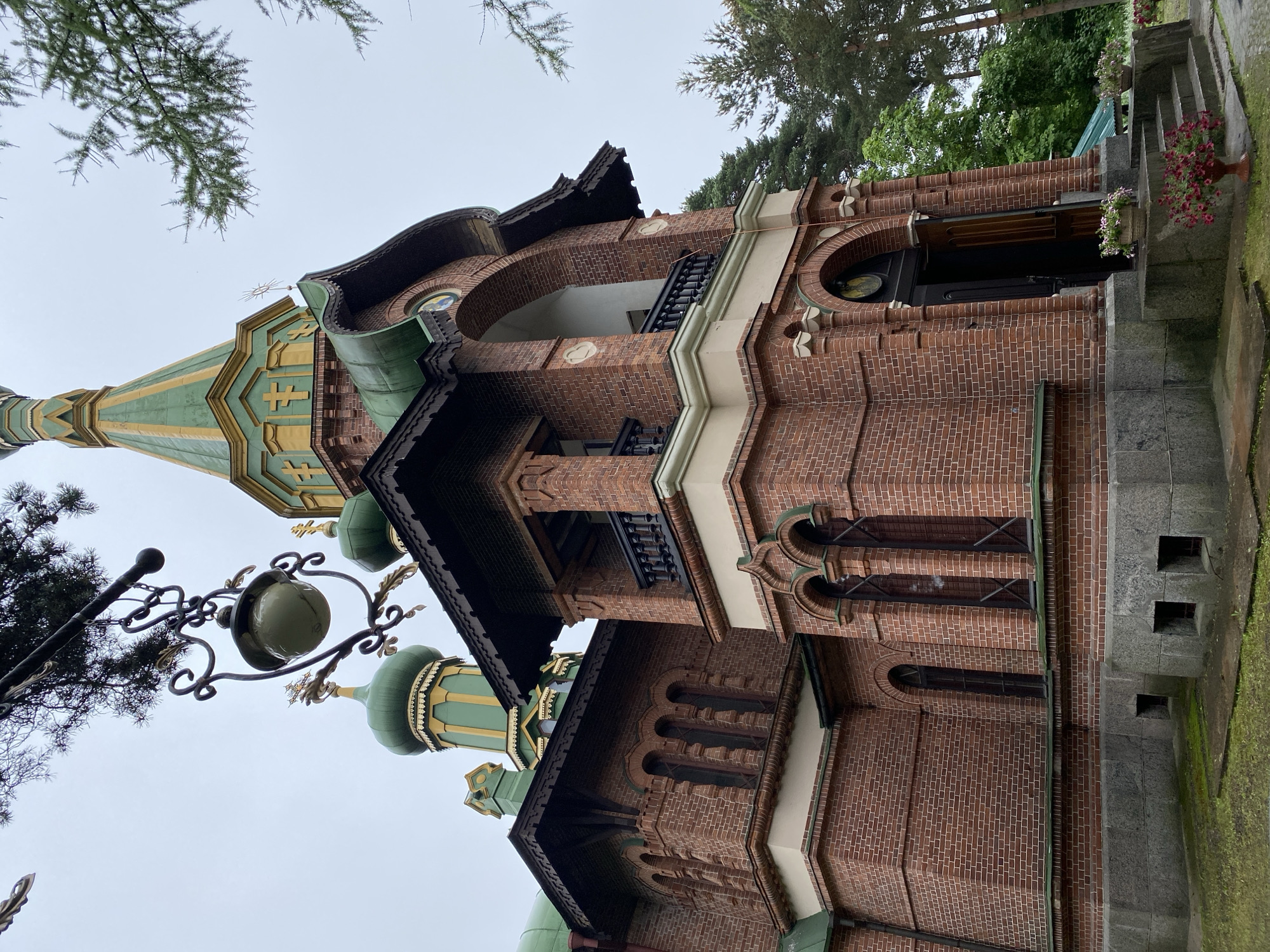 The image depicts the Church of the Assumption of the Blessed Virgin Mary in Priozersk, Russia. The structure is primarily made of red brick with green domes and golden accents. The foreground features stone steps leading to a wooden entrance, adorned with potted flowers. A decorative lantern hangs from a wrought-iron fixture above and to the left of the building. Lush green trees surround the church. The sky is overcast.