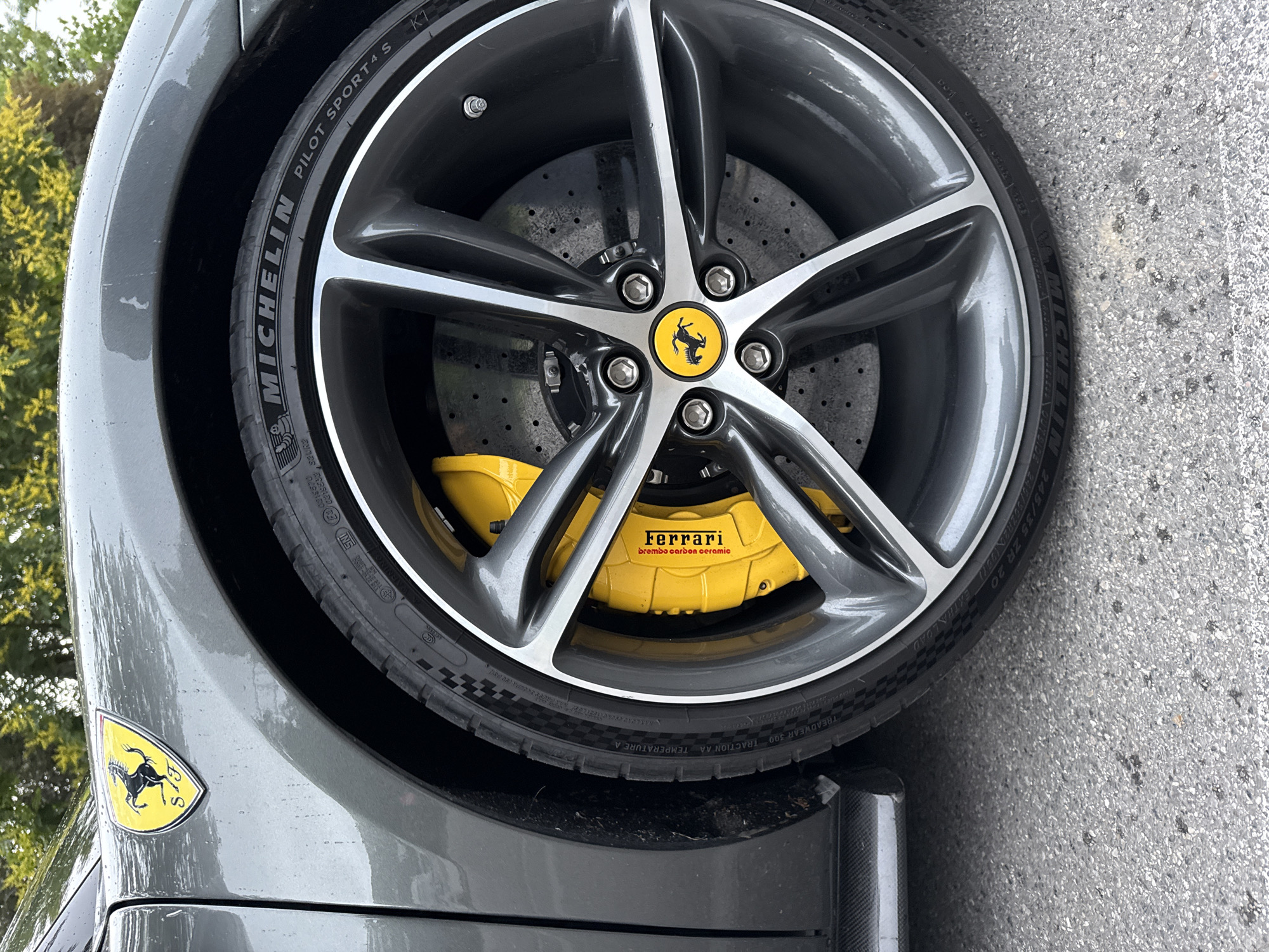 This is a close-up, high-angle outdoor shot of the front wheel and fender of a grey Ferrari. The car has a yellow Ferrari emblem on the fender and yellow brake calipers with "Ferrari" and "Brembo carbon ceramic" written on them. The wheel is dark grey with silver accents and a yellow Ferrari center cap. The tire is a Michelin Pilot Sport 4S. The car is parked on asphalt. The background is blurred and shows trees and greenery, suggesting a park or a residential area. The image was taken in Vienna, Austria, on a slightly wet day, as evidenced by water droplets on the car's surface. There are no people or animals visible in the image.